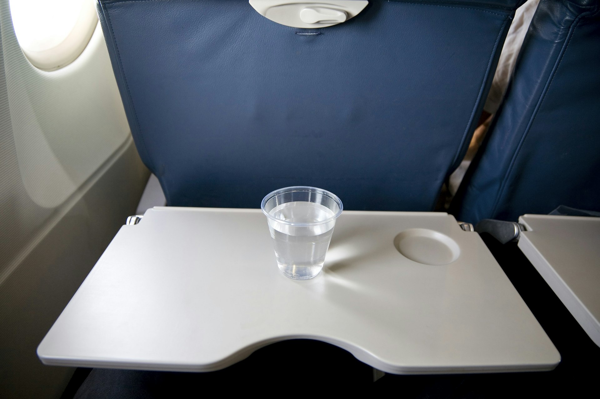 A plastic glass of water sits on a table track on the back of an airplane seat.