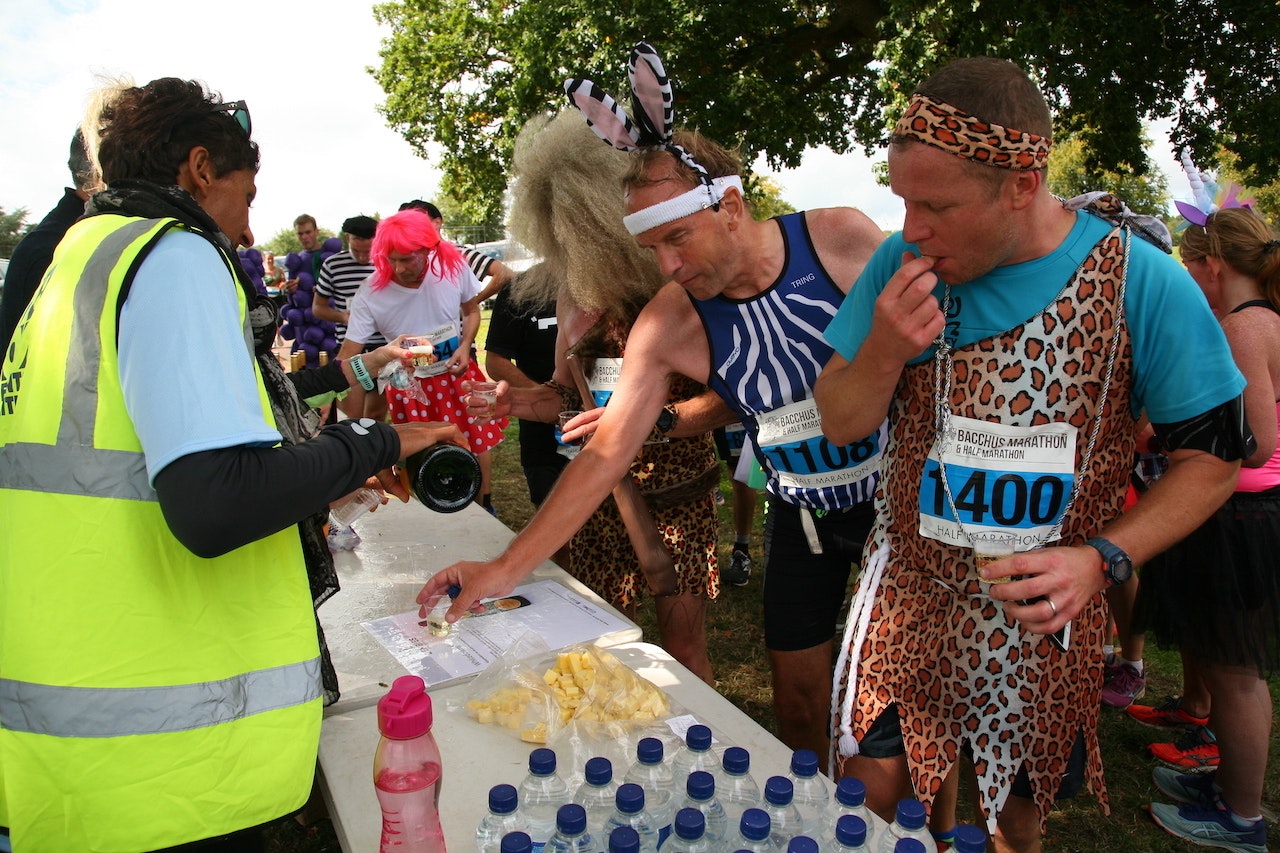 Runners in fancy dress have stopped at a refreshment station. A woman in a hi-vis tabard is pouring wine for them to taste. Several bottles of water remain unopened.