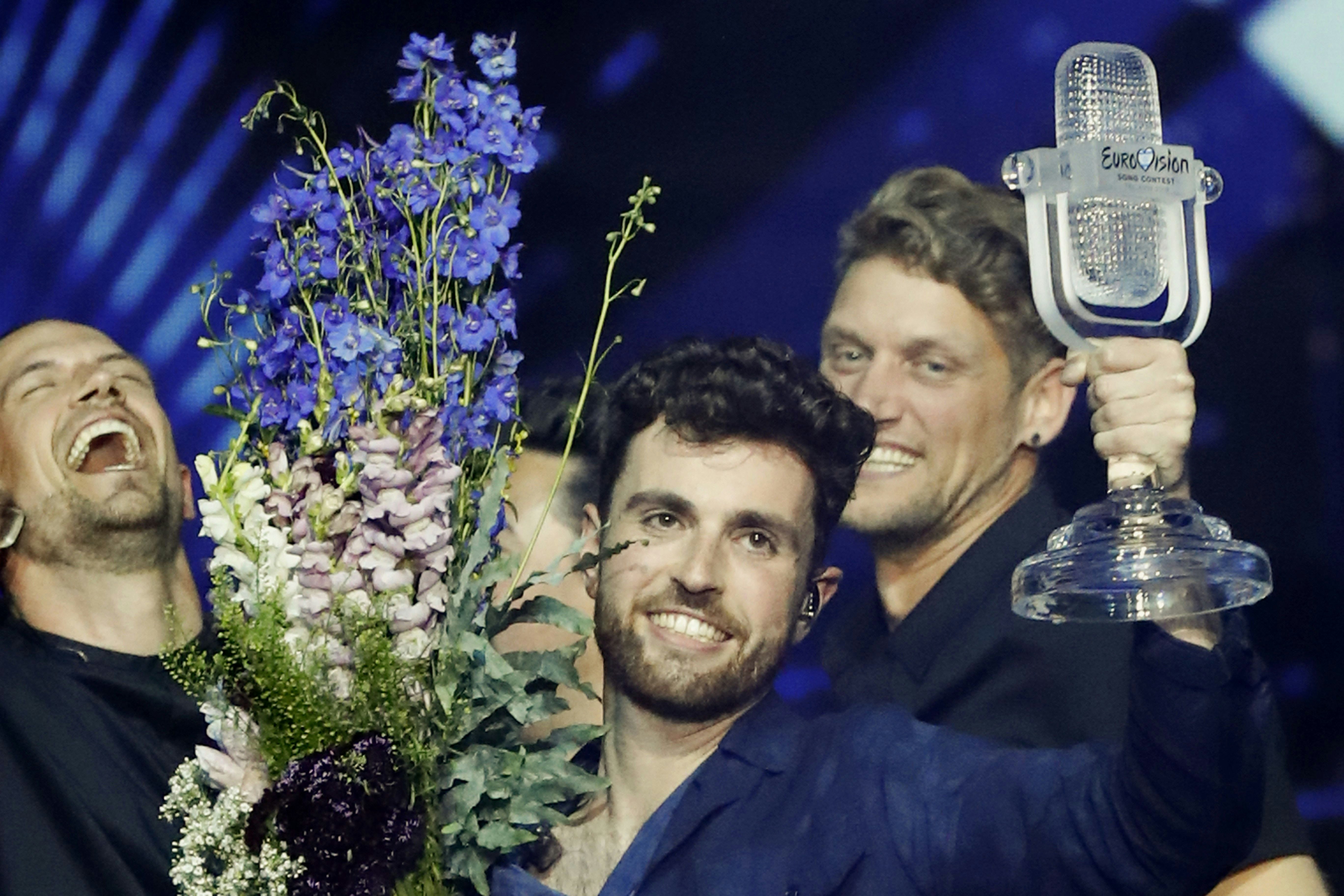Winner of Eurovision 2019, Duncan Laurence holds up the glass microphone trophy in one hand and a bouquet of flowers in the other, as his entourage smile and laugh behind.