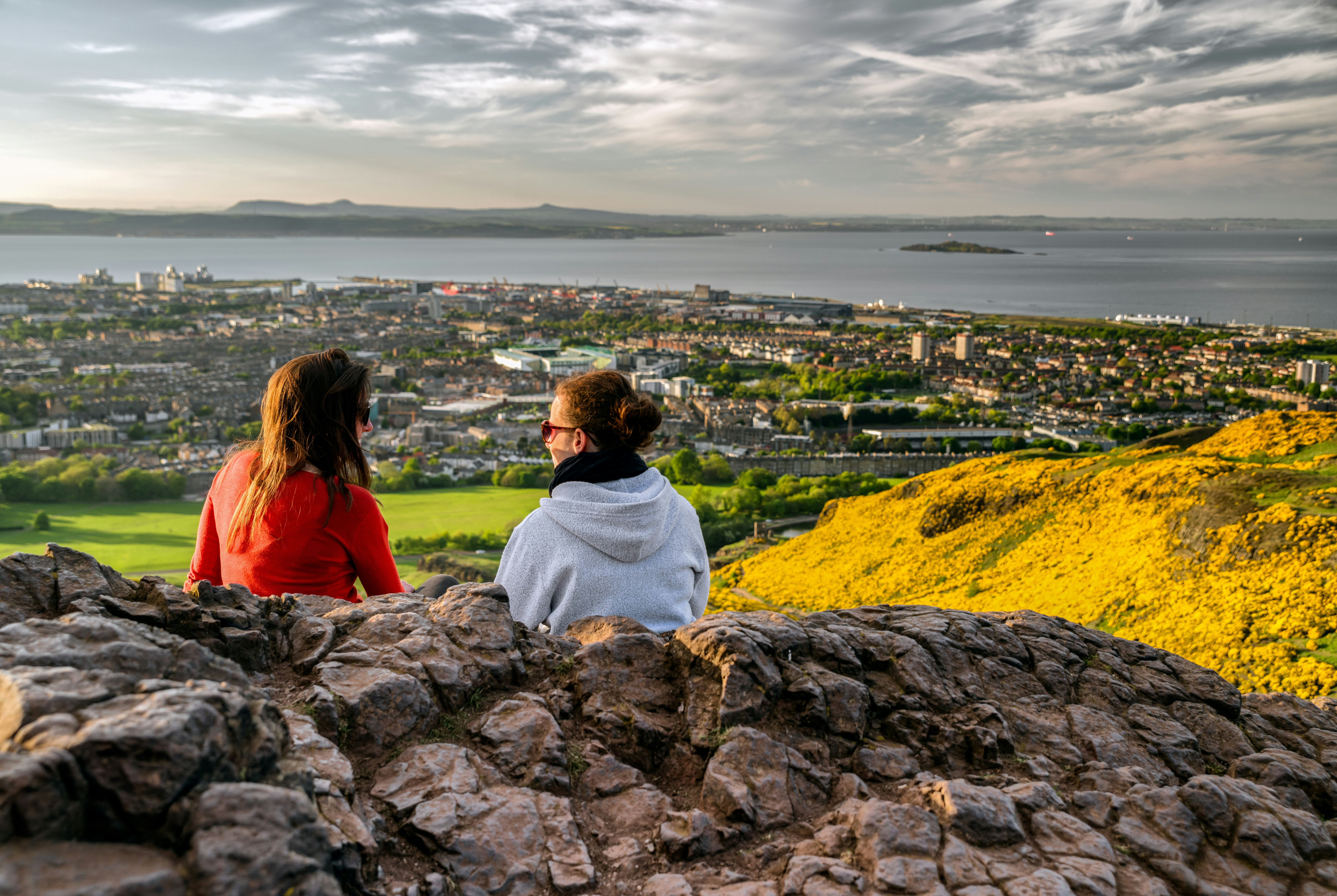 Two girls sitting on the hill of Arthur's Seat overlooking Edinburgh. The hills are covered in yellow wild flowers and the sky is cloudy and moody. 