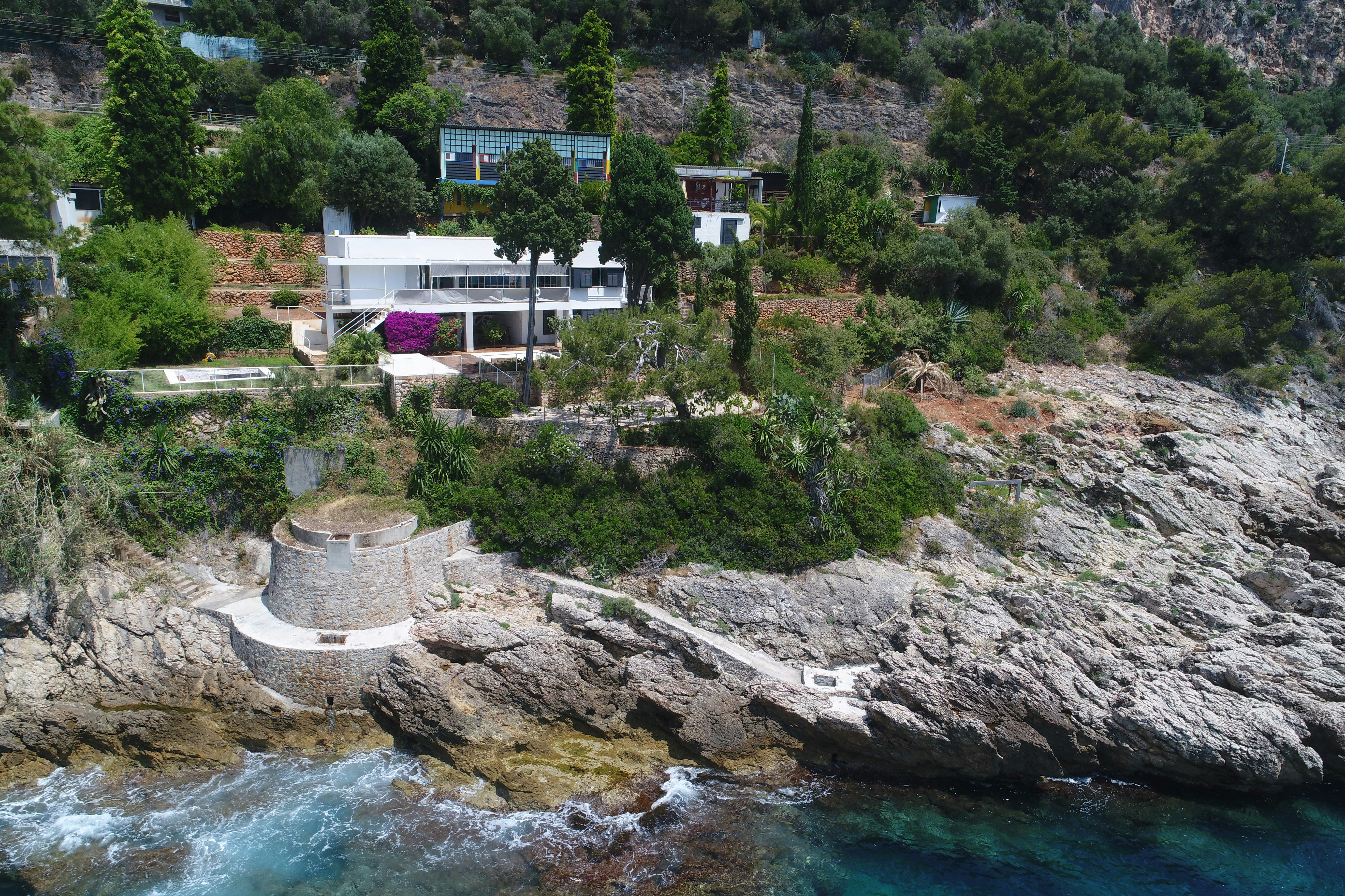 A striking white modernist villa, partially obscured by large trees, set on the rocky Cap Moderne, Roquebrune-Cap Martin, France