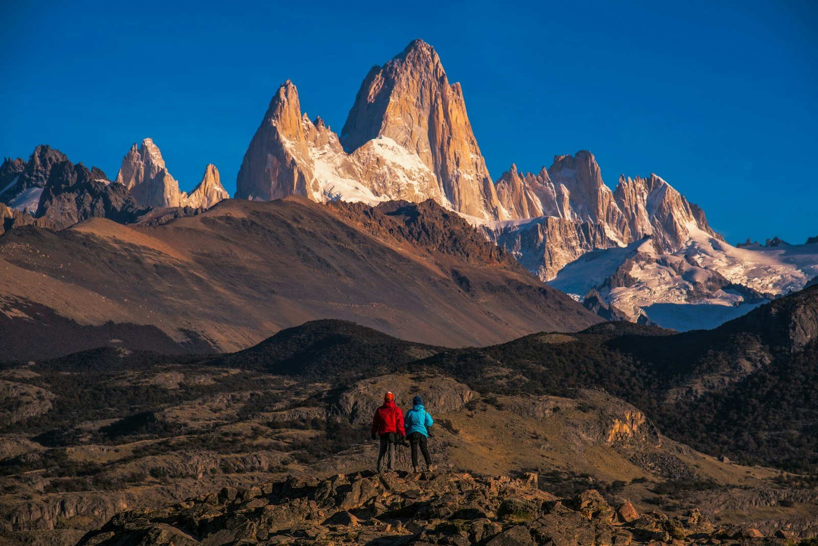 A couple stand with their backs to the camera and look over a parched landscape and up to the highest summits of Los Glaciers National Park.