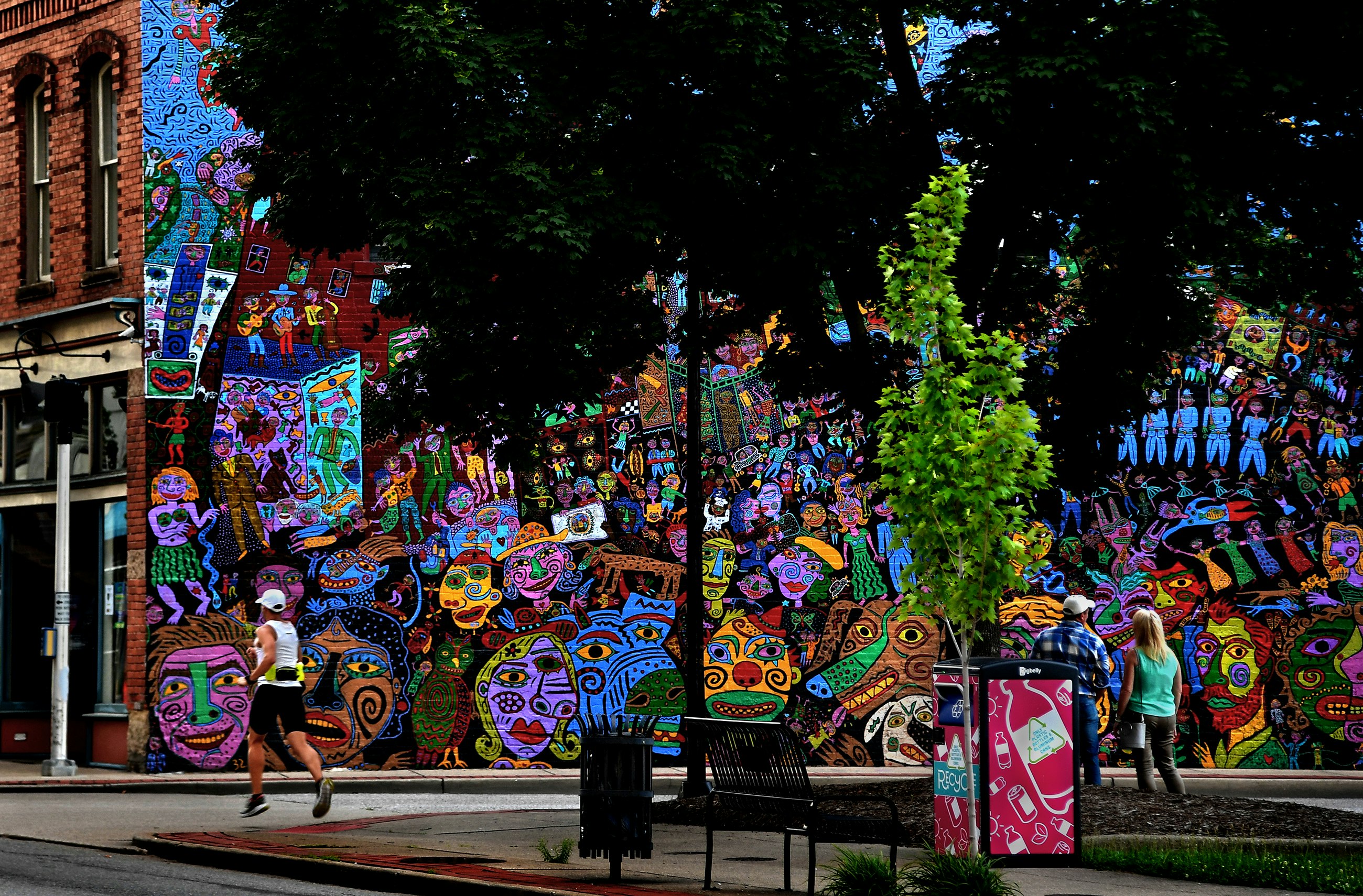 A colorful mural showing abstract faces adorns the side of a historic brick building, partially obscured by trees. A man and a woman stand and admire the mural while a jogger runs by. A trashcan and park bench are in the foreground
