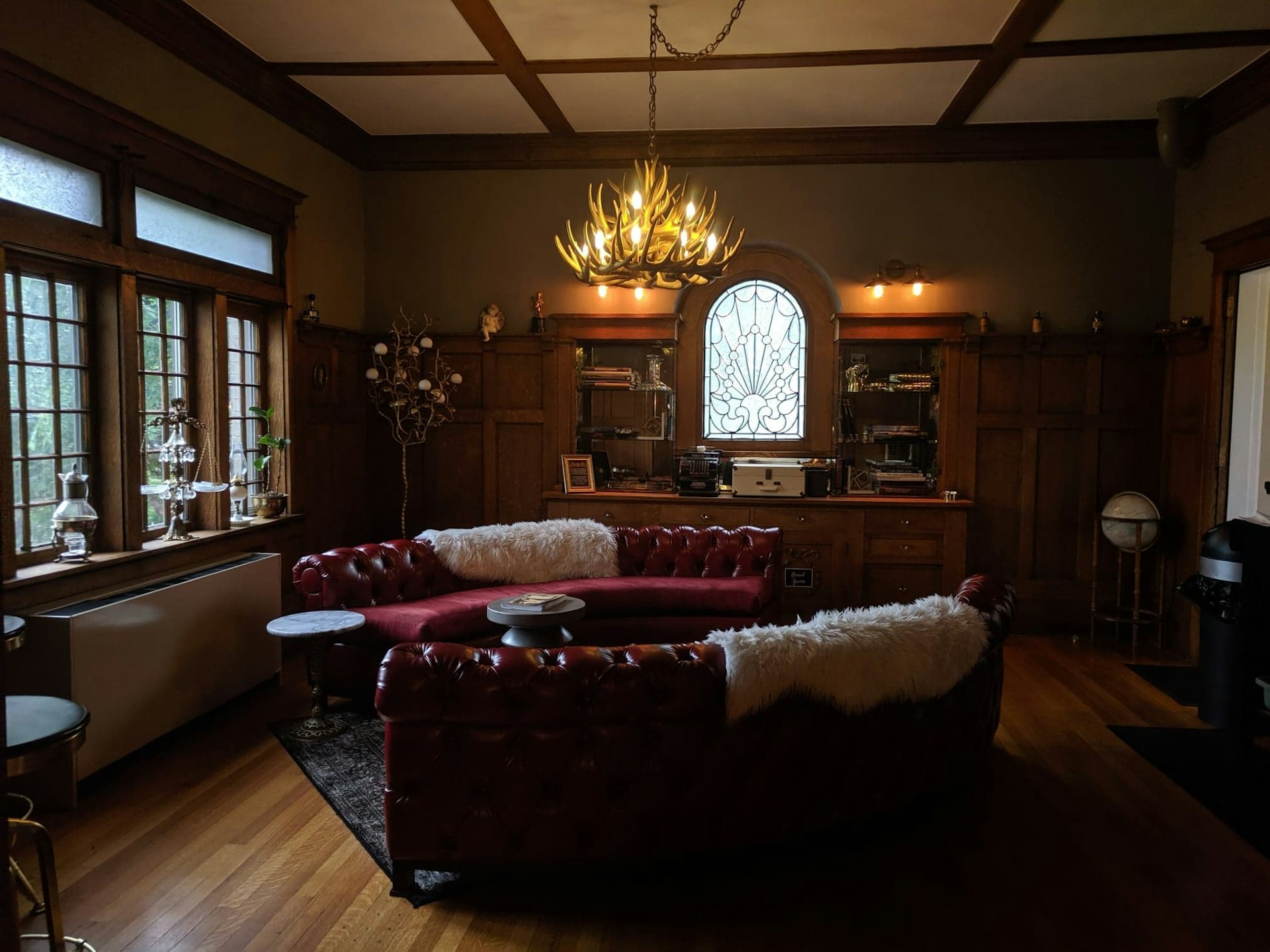 A seating area at Ember Hostel Denver is in an elegant wood paneled room with large glass windows, including one made of clear stained glass surrounded by a built-in credenza. There are two semicircular red leather Chesterfield sofas with white faux sheep skins draped over them. Above is a chandelier made of antlers. 