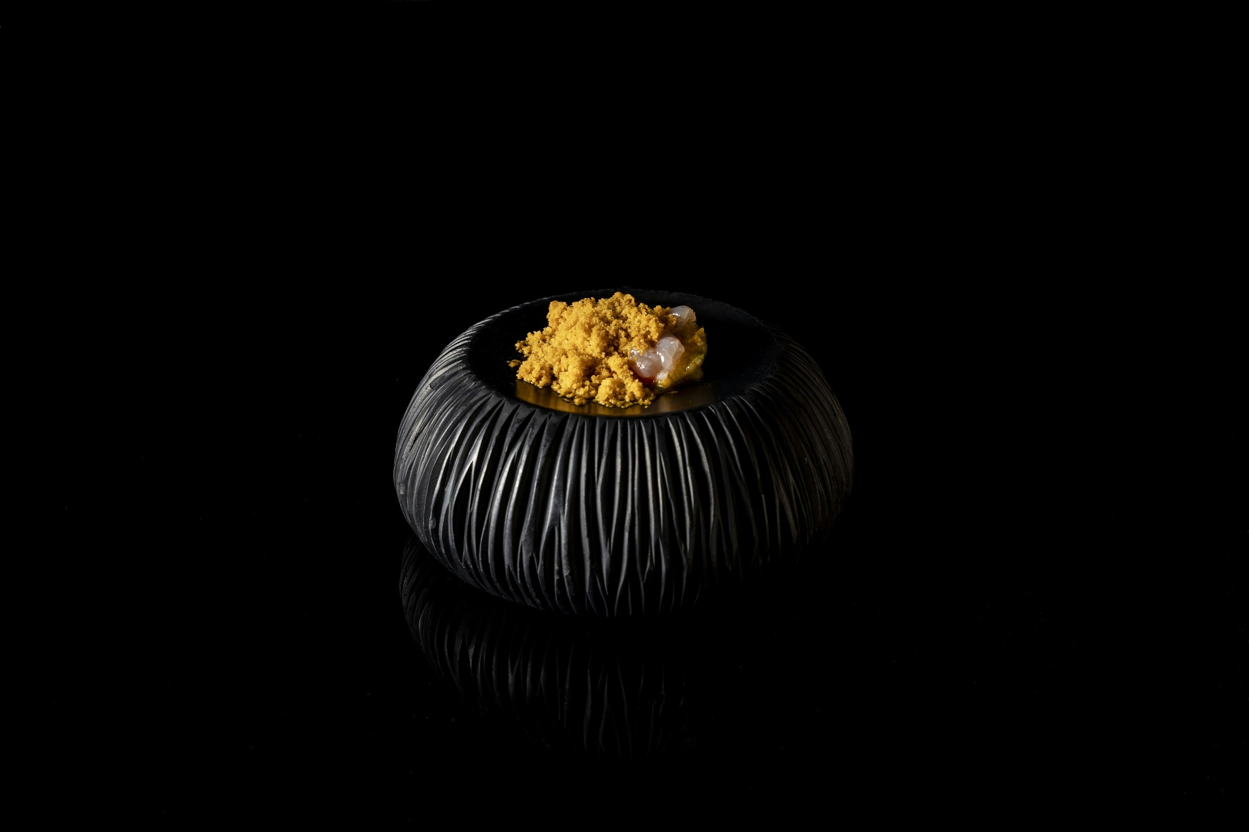 Ett tjockt svart fat, med tillhuggna sidor som liknar sidorna på en vulkan, rymmer en delikat servering av en gurkmejafärgad maträtt.