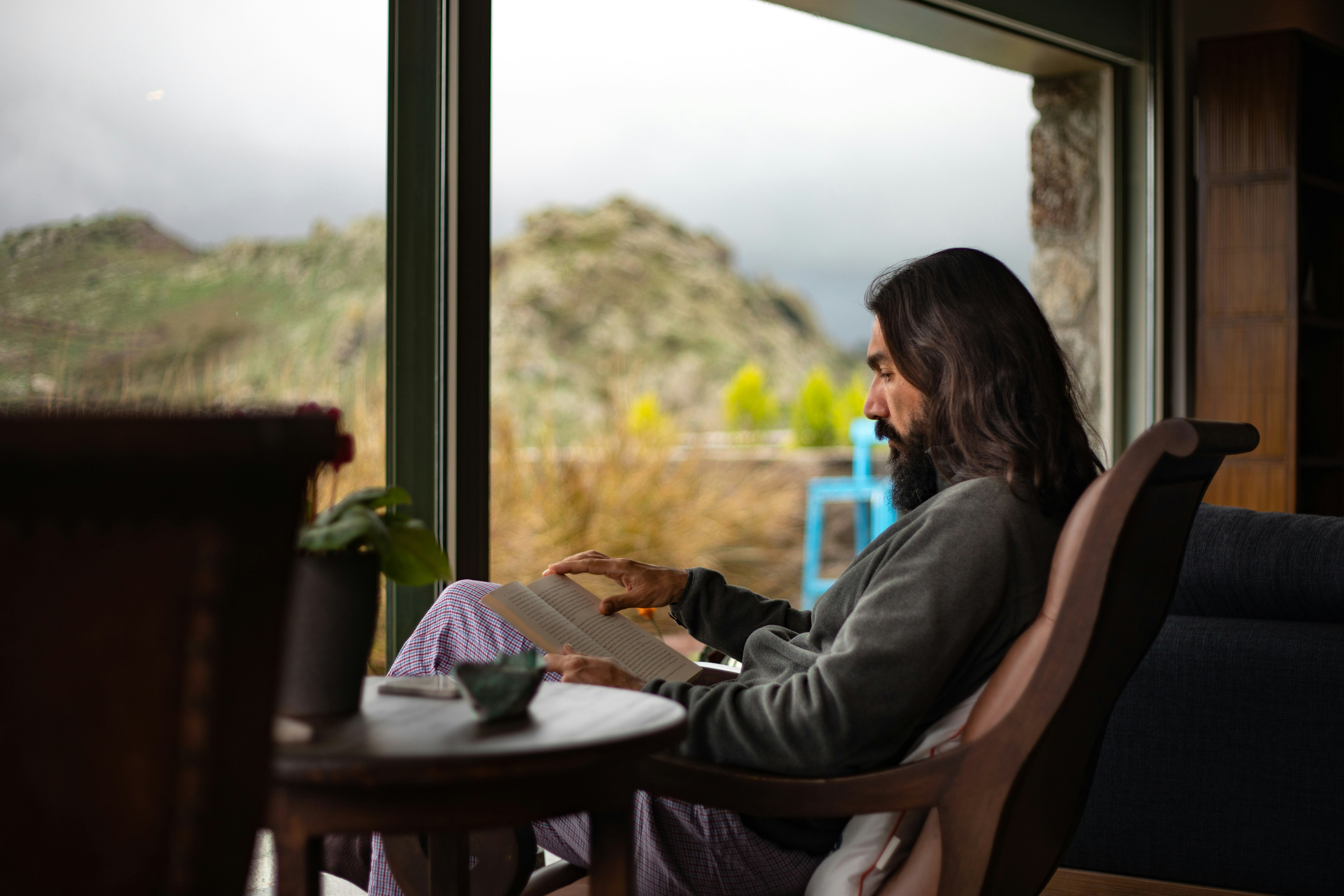 A man with long hair and a beard in a grey fleece pullover and blue and red patterned pajama pants reads a book in a cozy living room with a view of a rocky hill outside the picture window