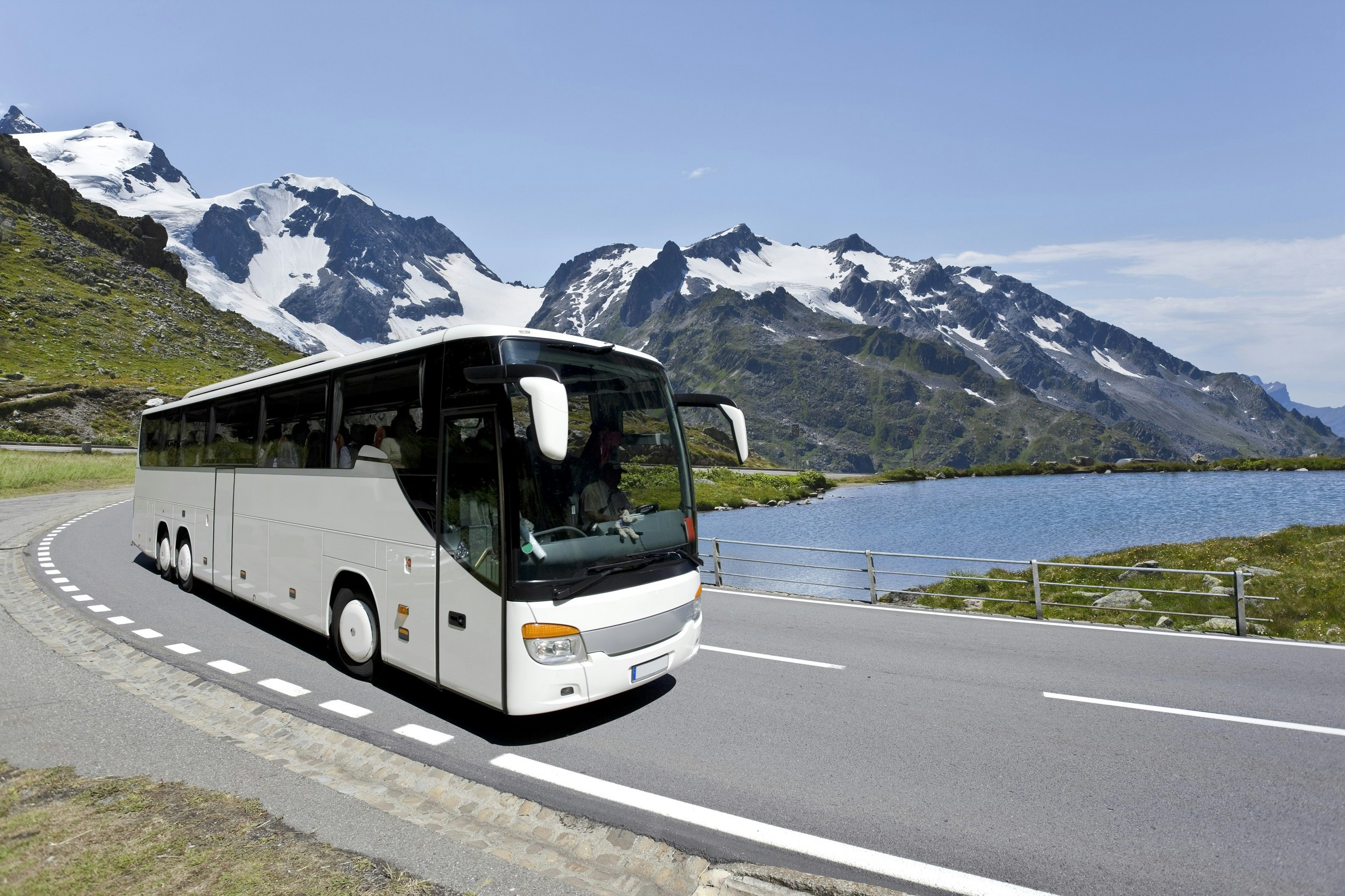 A large white coach passes a lake, with snow-topped mountains in the background