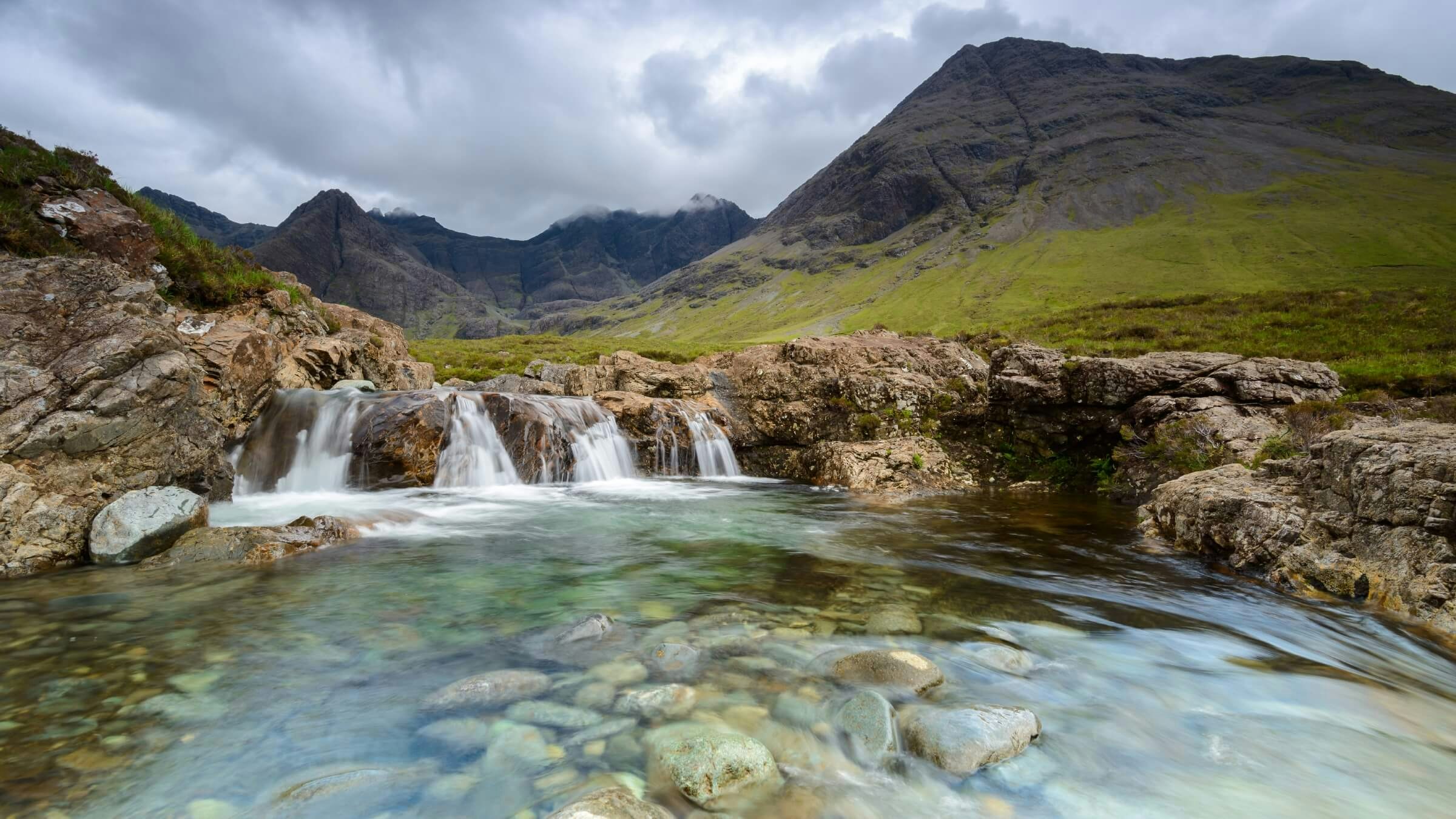 A pool of clear water with stones visible at the bottom. A small stream flows into it and mountains loom in the background.