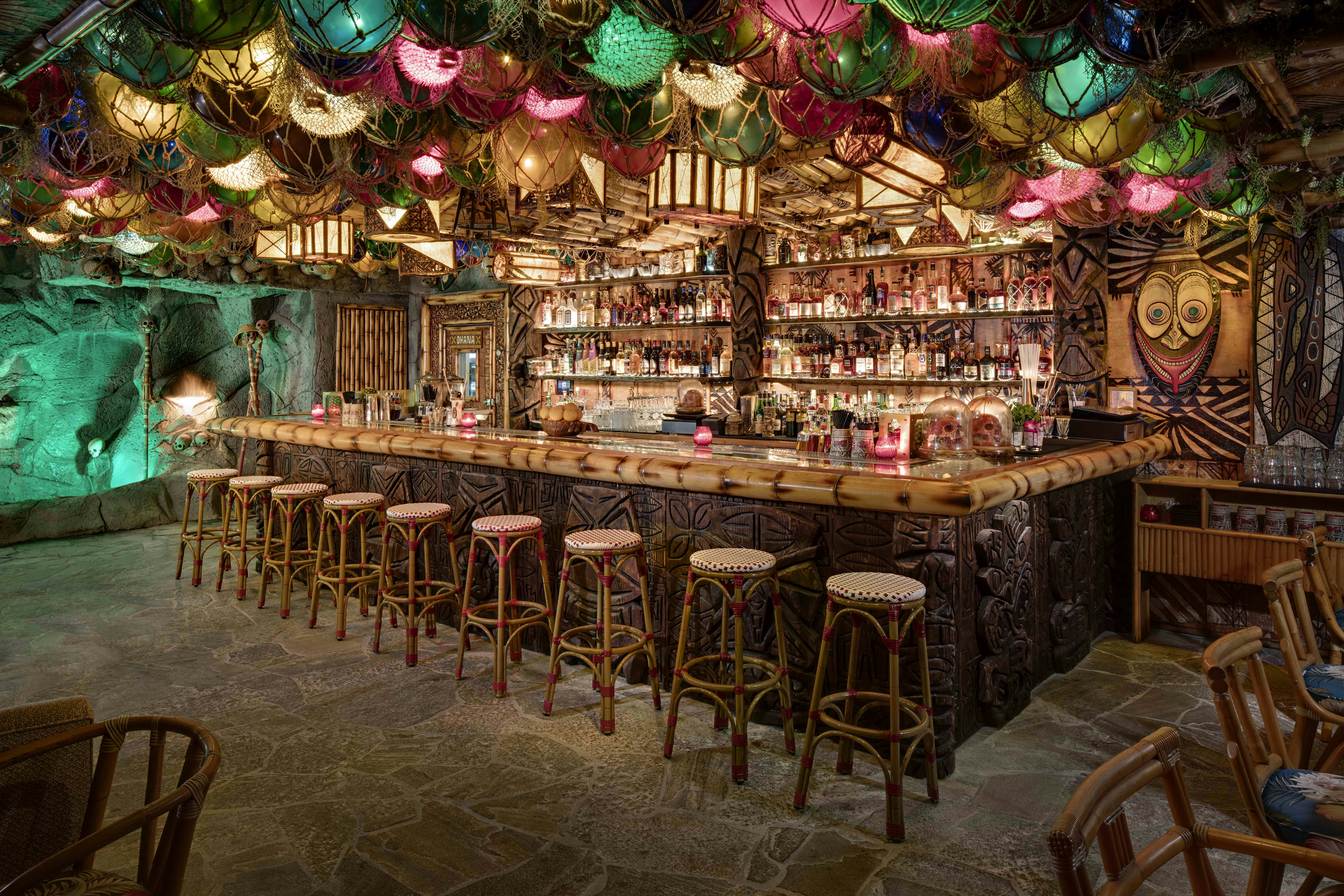 Ratan stools line up in front a bar whose front and sides are made up of dark wood carved tiki motifs and the top is like a large piece of bamboo. Colorful glass fishing floats in sisal nets hang from the ceiling. In the left corner is a faux cave wall decorated with skulls and a pith helmet