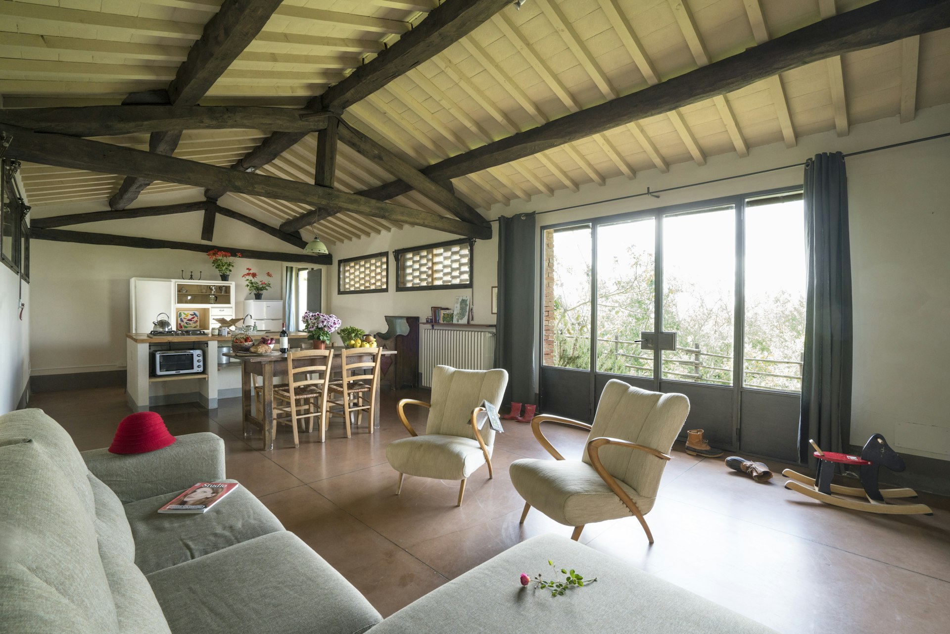 A spacious, rustic great room at Fattoria Barbialla Nuova features midcentury modern minimalist furniture, rustic farmhouse beams overhead, a child's wooden rocking horse, and a red hat sitting on the sofa