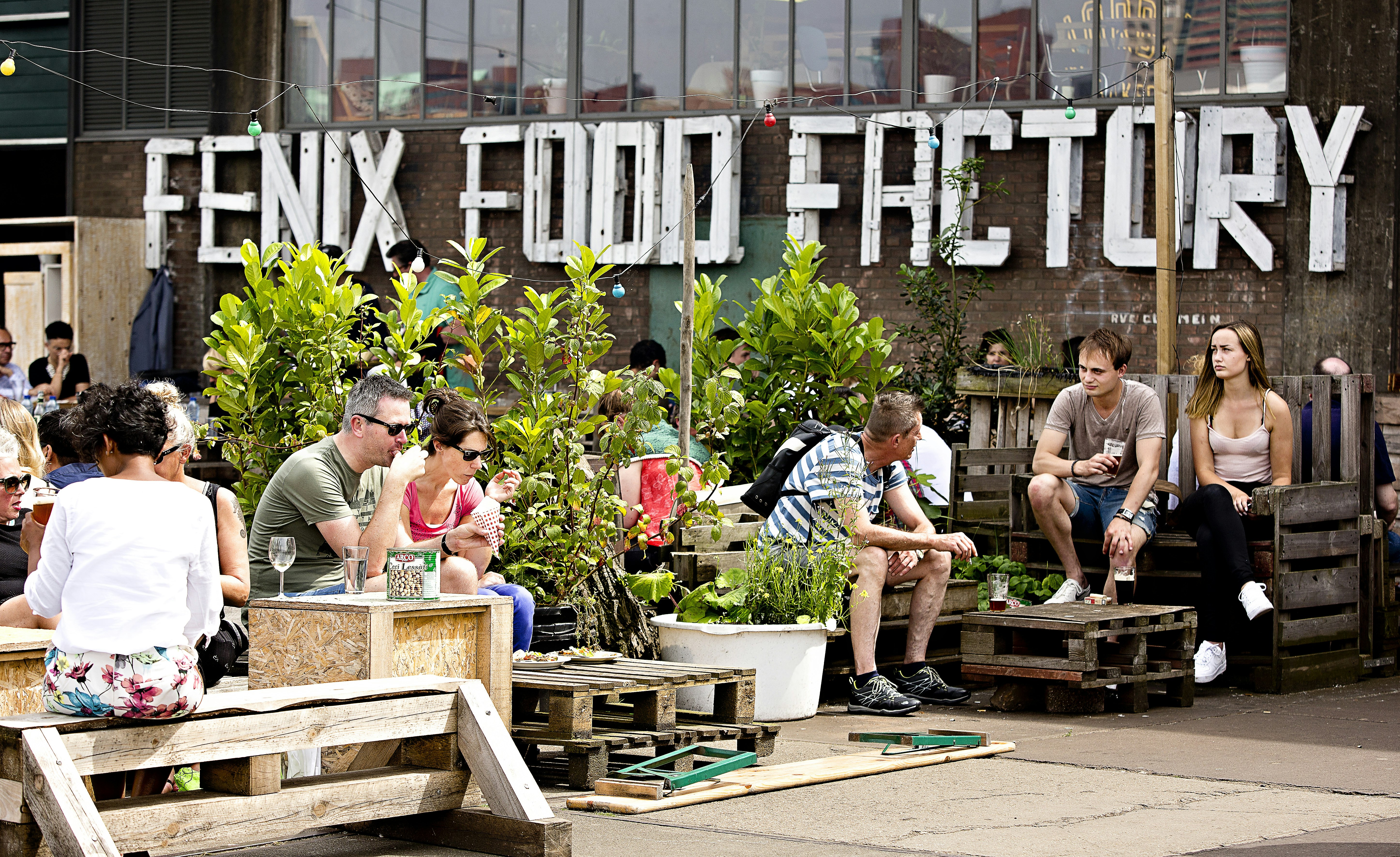 Many people are sitting on makeshift wooden-pallet furniture outside converted warehouse Fenix Food Factory in Rotterdam on a sunny day.