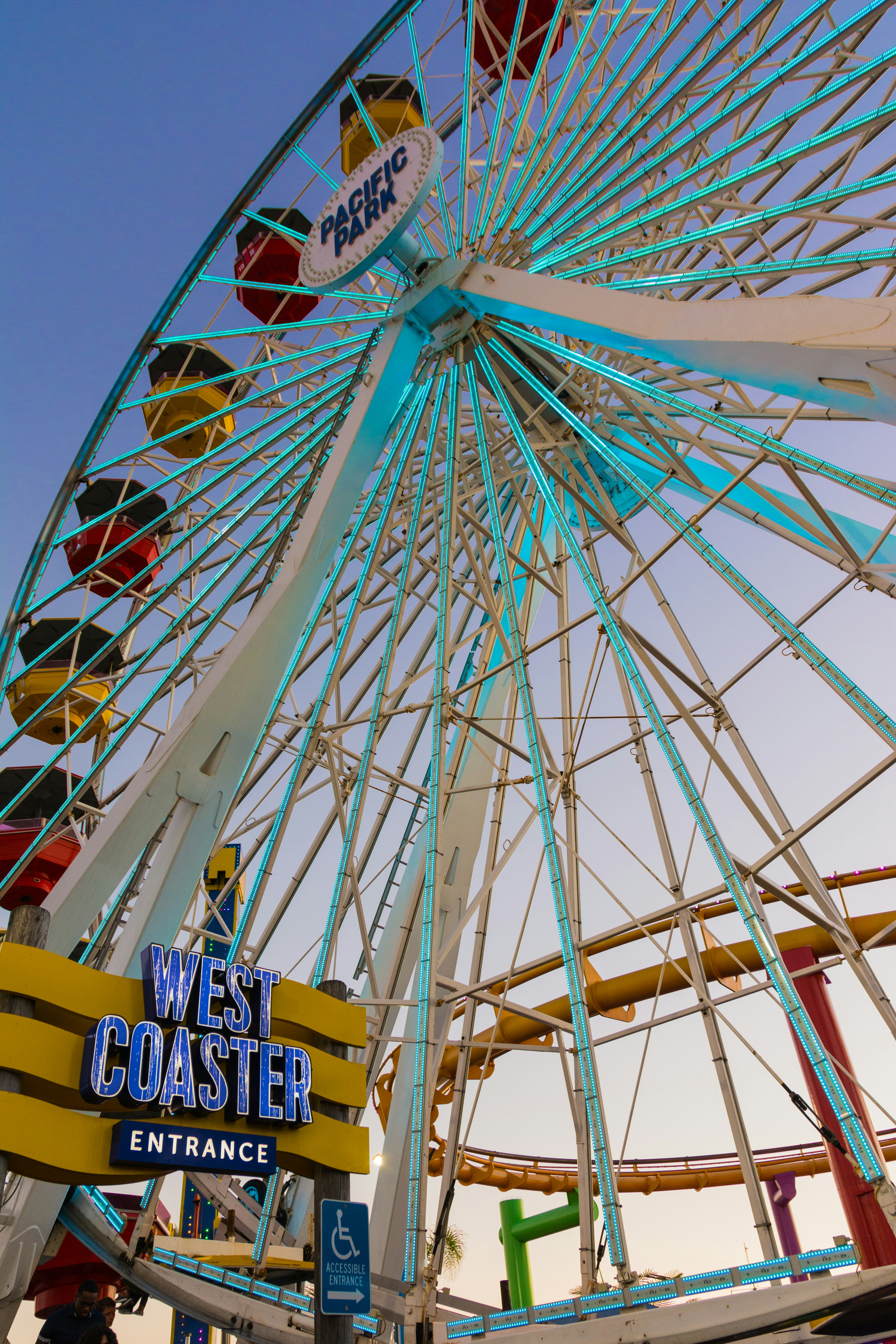 A close-up view looking up at the ferris wheel on Santa Monica Pier. An illuminated sign says 'West Coaster Entrance'.