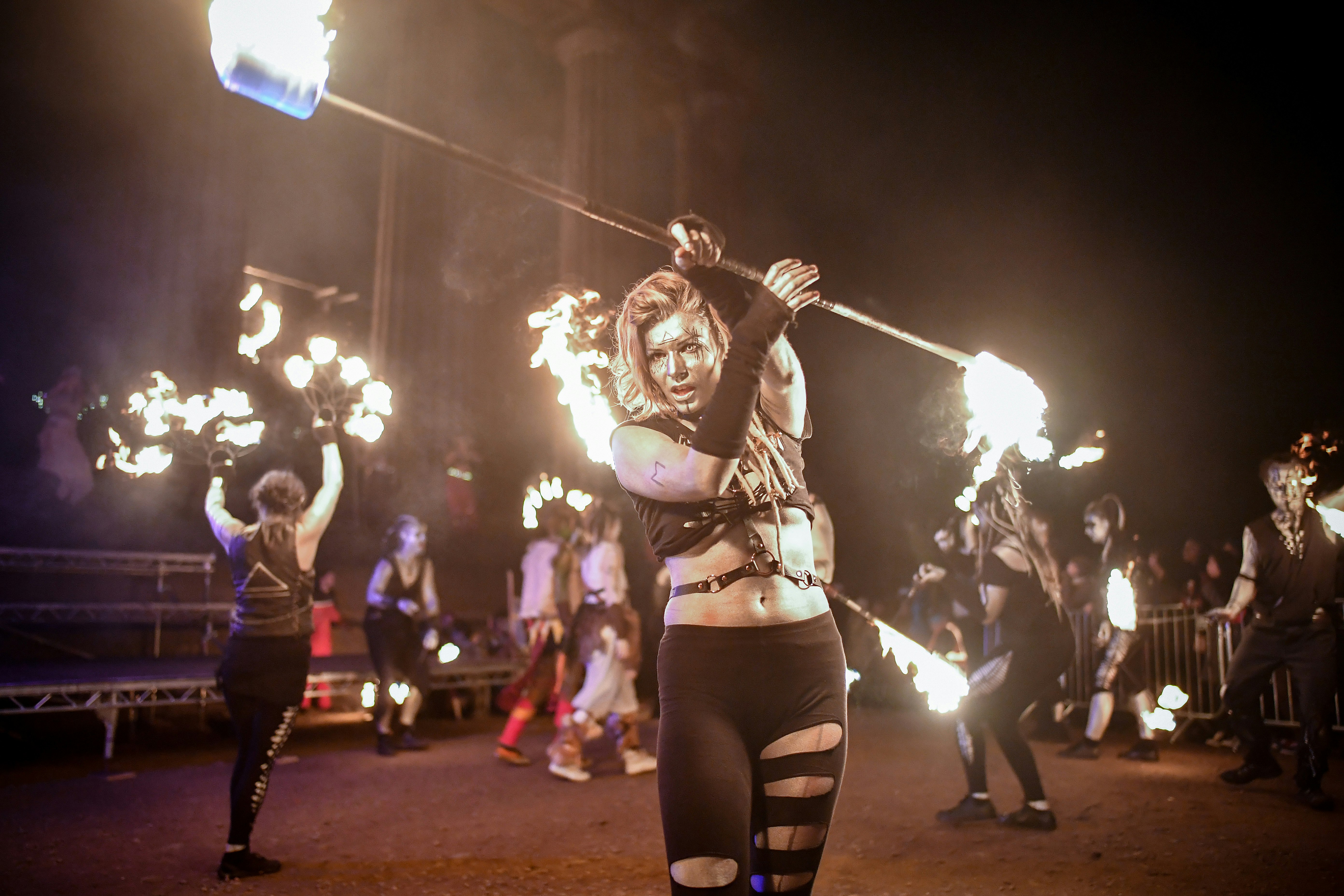 A woman in black, artfully ripped tights, black arm warmers, and a leather harness draped over her are midriff twirls a staff that's flaming at both ends as other fire dancers perform in the background at Carlton Hill on Samhuinn in Edinburgh, Scotland