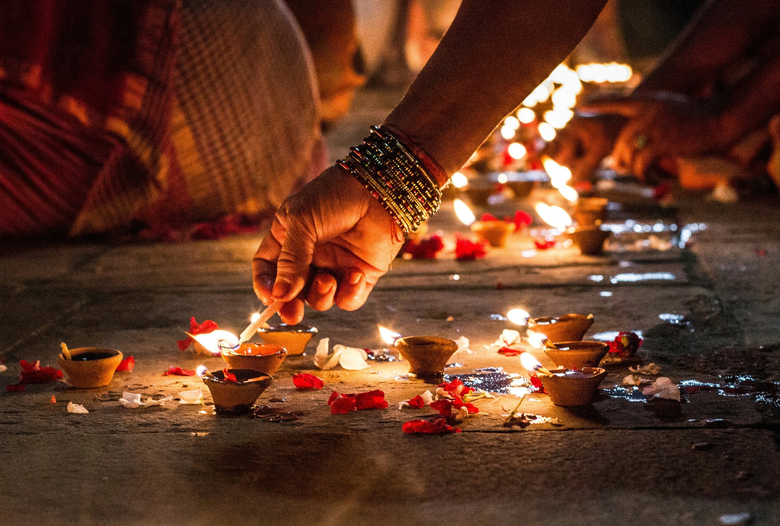 An arm with many bracelets reaches down with a little candle in hand to light some small candles in tiny clay containers; in the distance are countless similar containers glowing with flame.