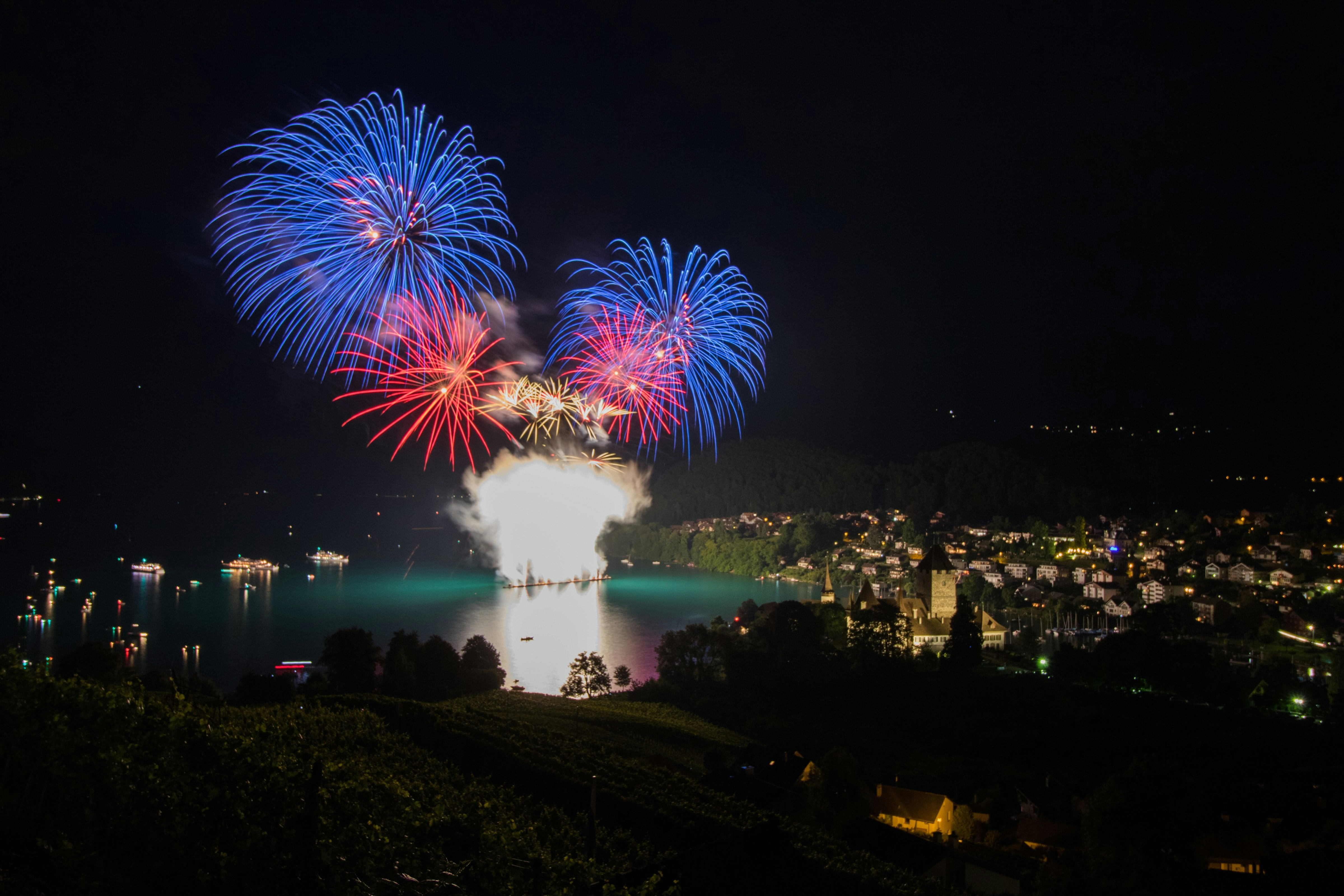 A boat in the middle of a blue lake fires colourful fireworks into the night sky. On the surrounding shores a number of houses are visible amongst greenery.