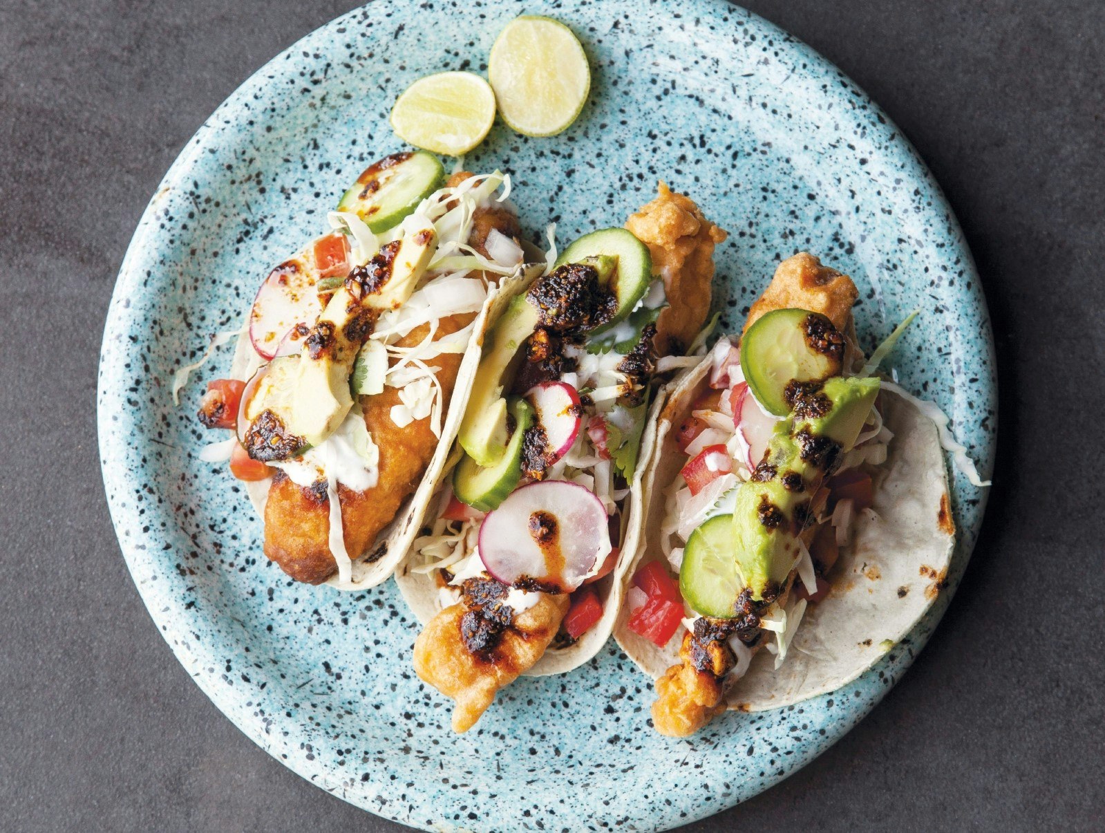 Three fish tacos: battered fish in corn tortillas with lettuce and sauce, served on a speckled plate.