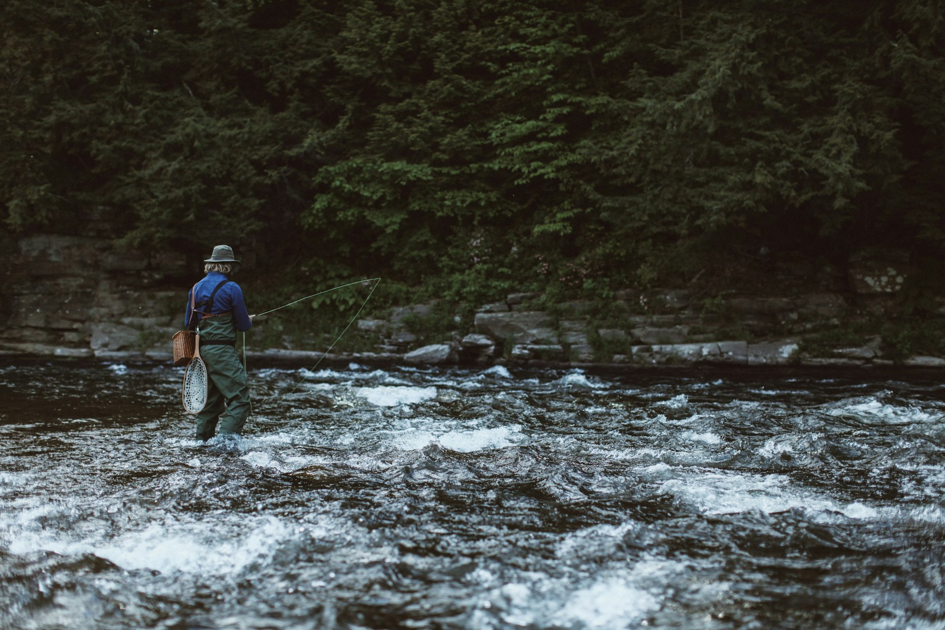 A man, wearing a cool hat and blue shirt, stands in the shallows of Beaverkill River, with rod in hand.