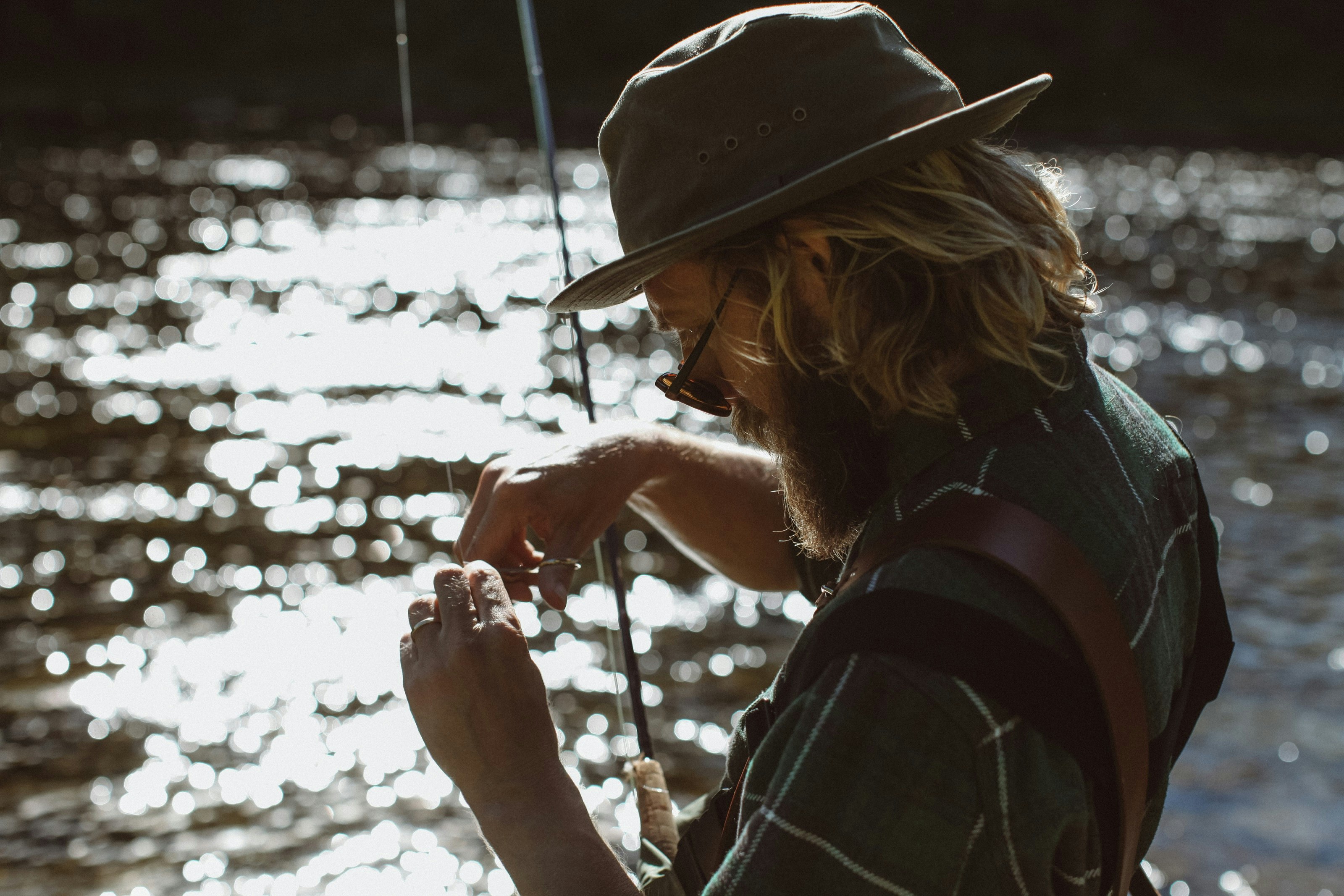 A man with long hair and sunglasses hooks a fly to his fishing rod while standing on the banks of a river. It's sunny and the sun is reflected in the waters of the river.