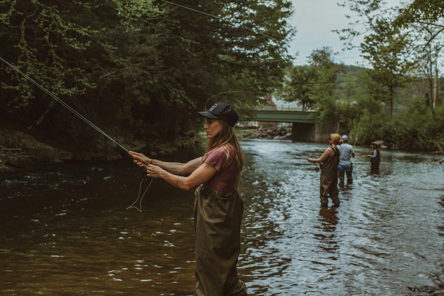 A small group of men and women stand in a river near the Livingston Manor Fly Fishing Club, practicing their cast.