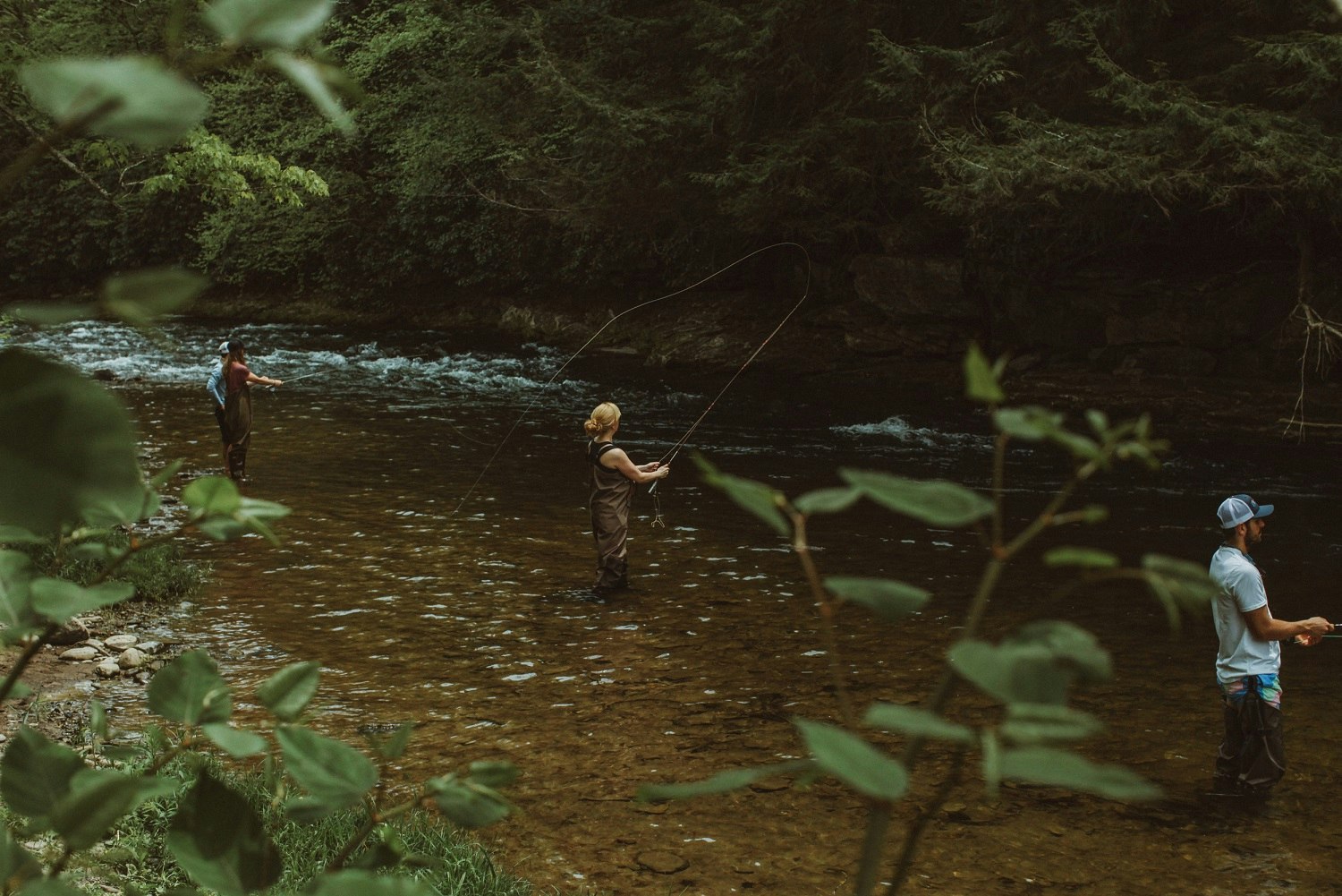An aerial view of some people standing in a river in the Catskills Mountains fly fishing. Leaves and other foliage frames the shot.