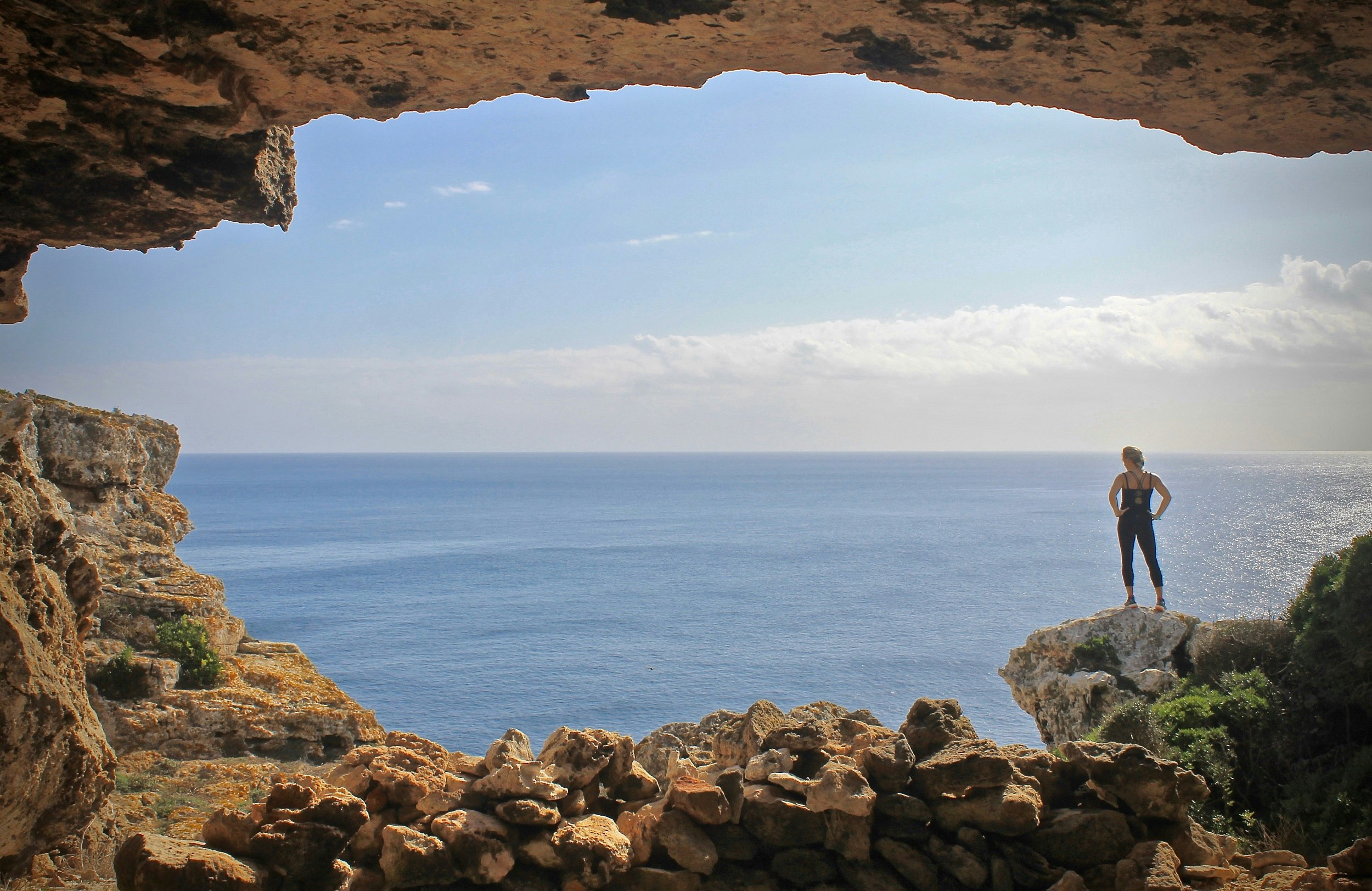A woman wearing an exercise kit stands on a rock on the outside of a large cave and overlooks the Mediterranean Sea.