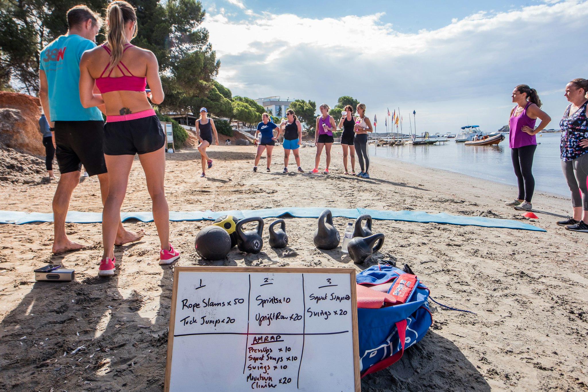 A group stand on a beach facing two fitness instructors; on the sand are mats, kettle bells and a white-board detailing the workout: rope slams x 50, tuck jumps x 20...