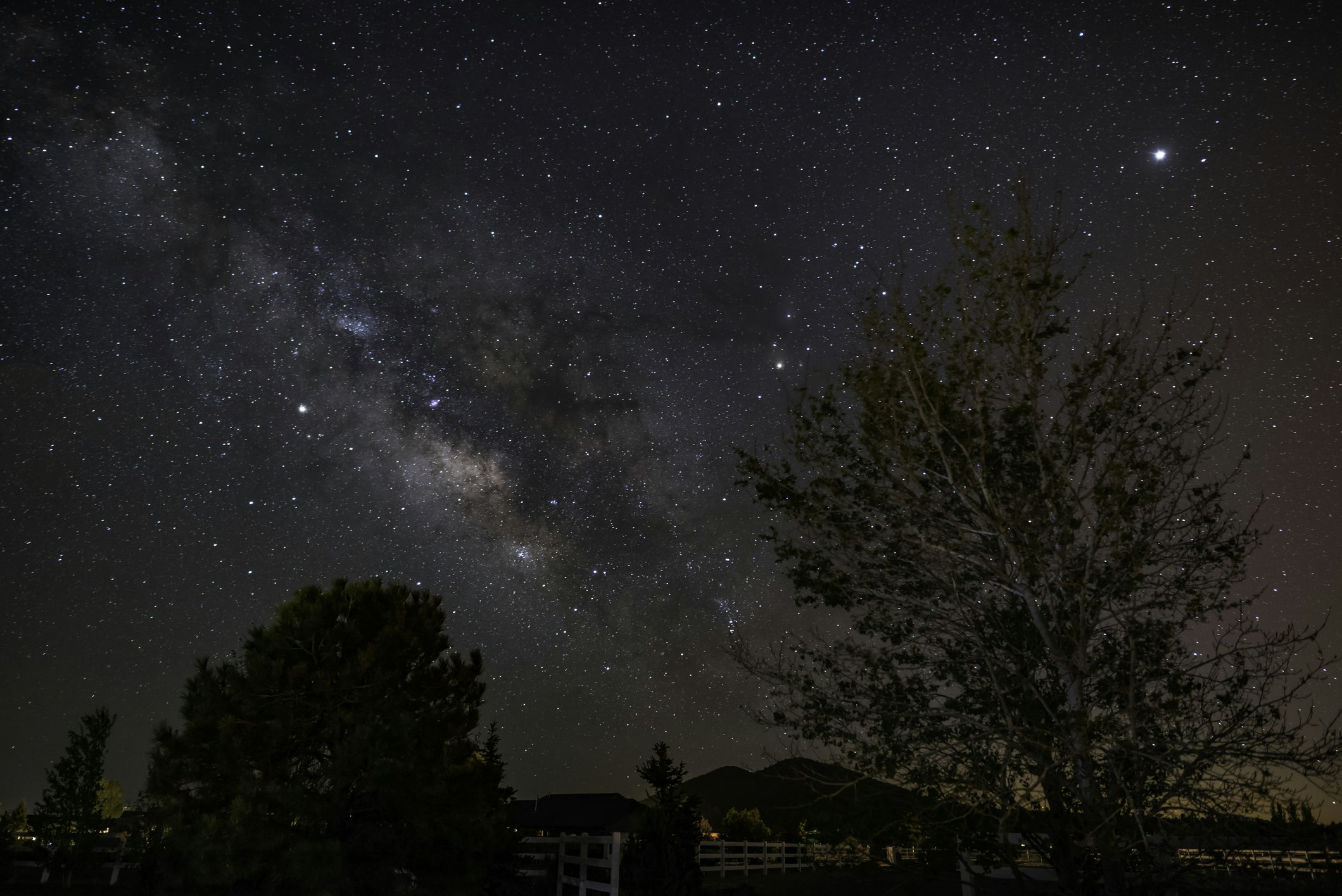 Stars and a small streak of the Milky Way appear over a low fence and medium tree in the foreground, with a small mountains and some other vegetation in the midground. The sky ranges from deep blue on the left to purplish red on the right, spangled with bright white stars and a low fringe of orange on the bottom right of the frame.