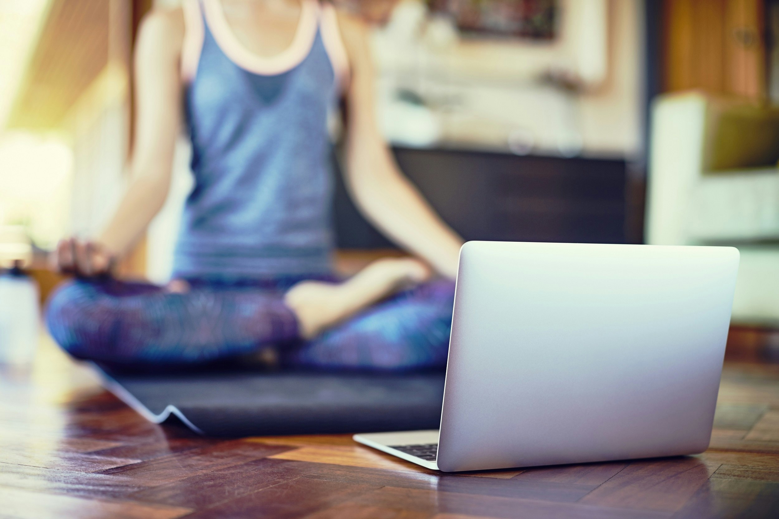 Low section of young woman practising yoga online from laptop at home; she is sitting in lotus position on an exercise mat. Focus is on a laptop in the living room.