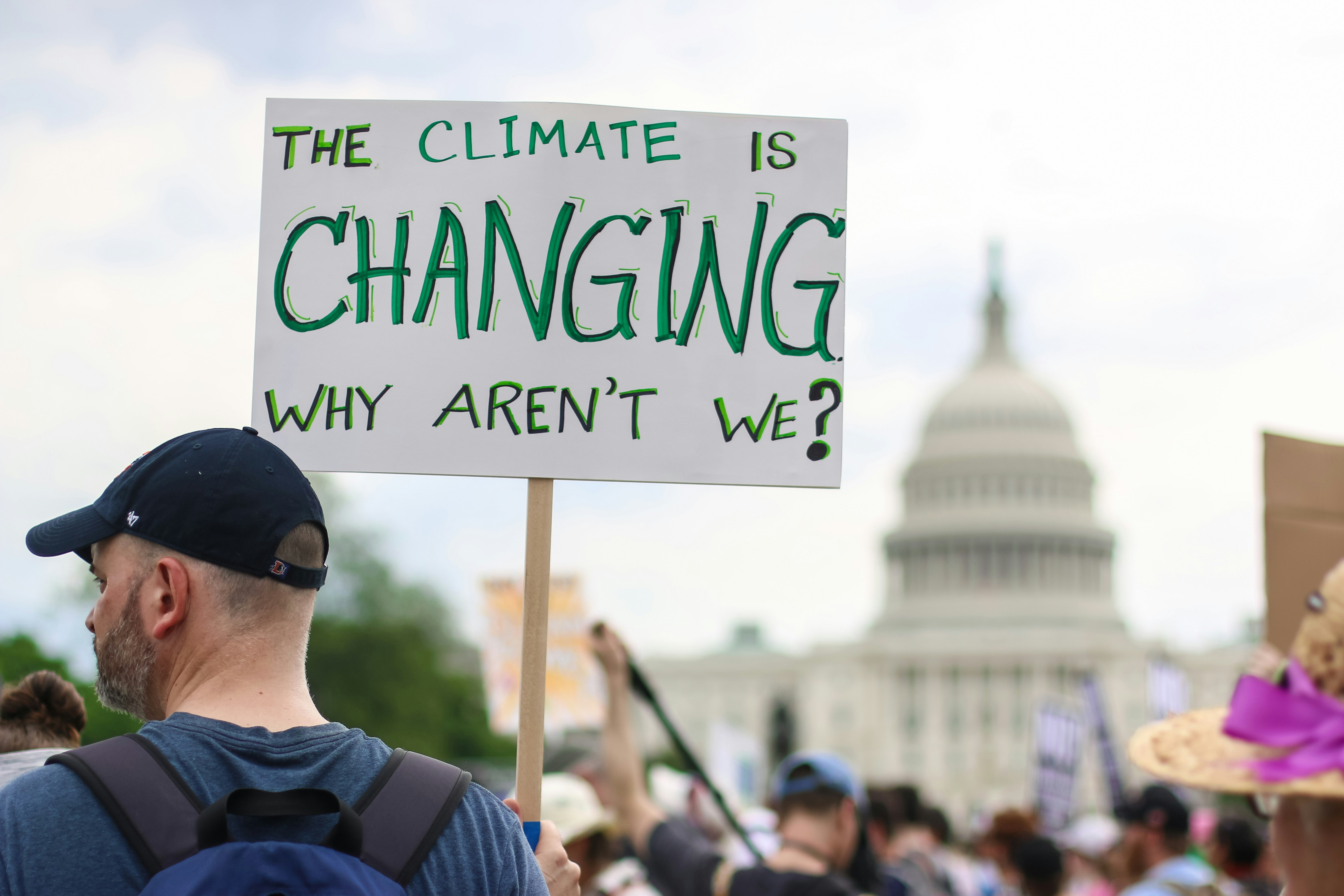 A man wearing a baseball cap holds a sign saying 'the climate is changing, why aren't we?'. The US Capital Building is in the background, out of focus.