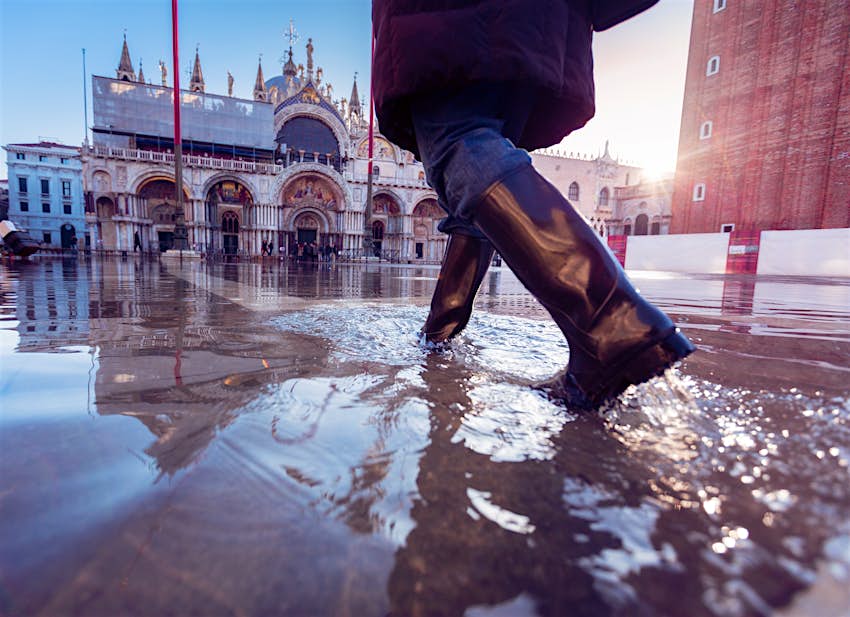 A person wearing rubber boots walks through flood waters while walking towards an ornate building at the edge of a large plaza