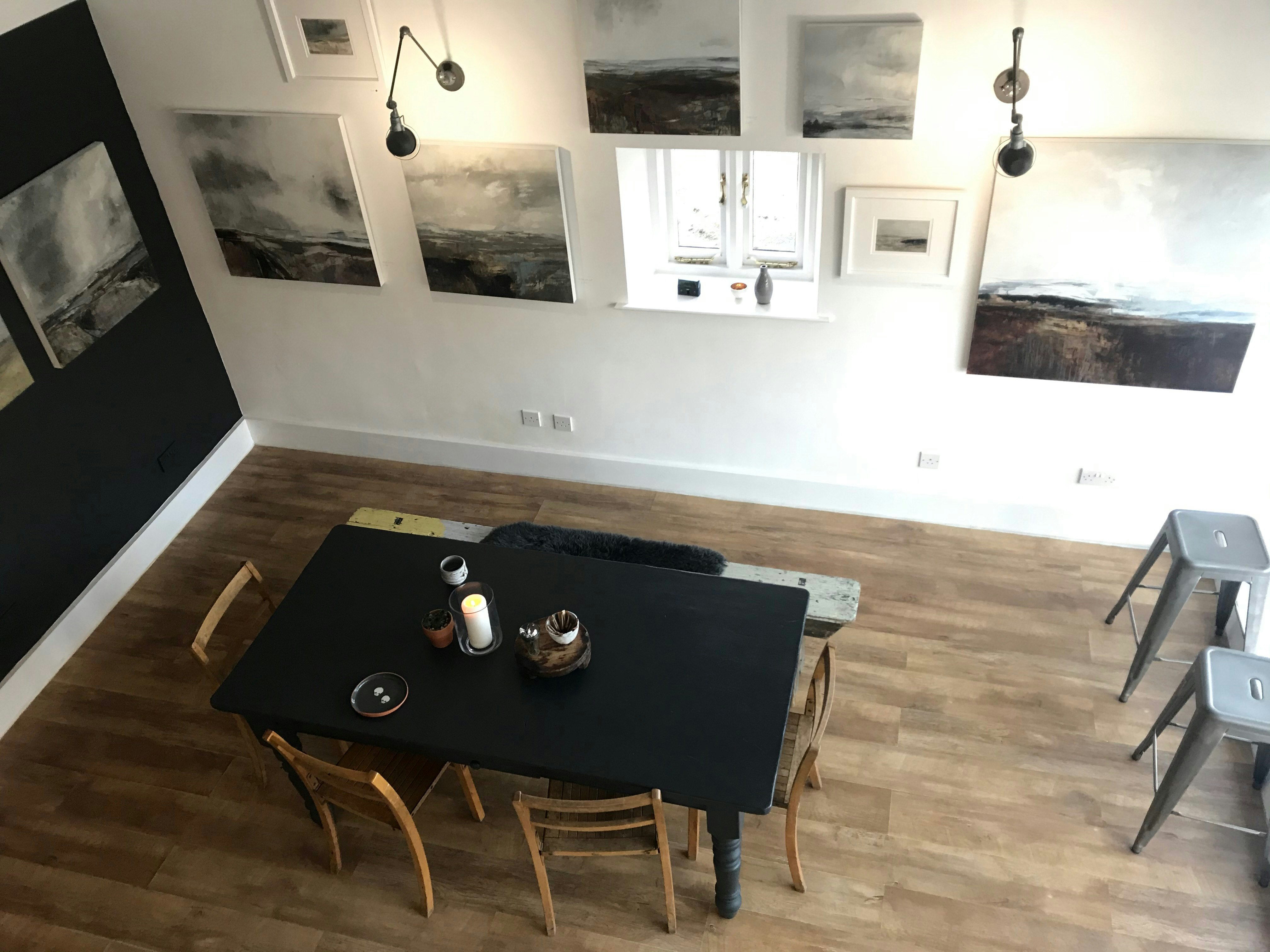 View looking down from a mezzanine level in a room in Gallery at Home, Usk. Black and white landscapes are hung on the monochrome walls while a minimalist dining table and chairs sits in the centre of the room.