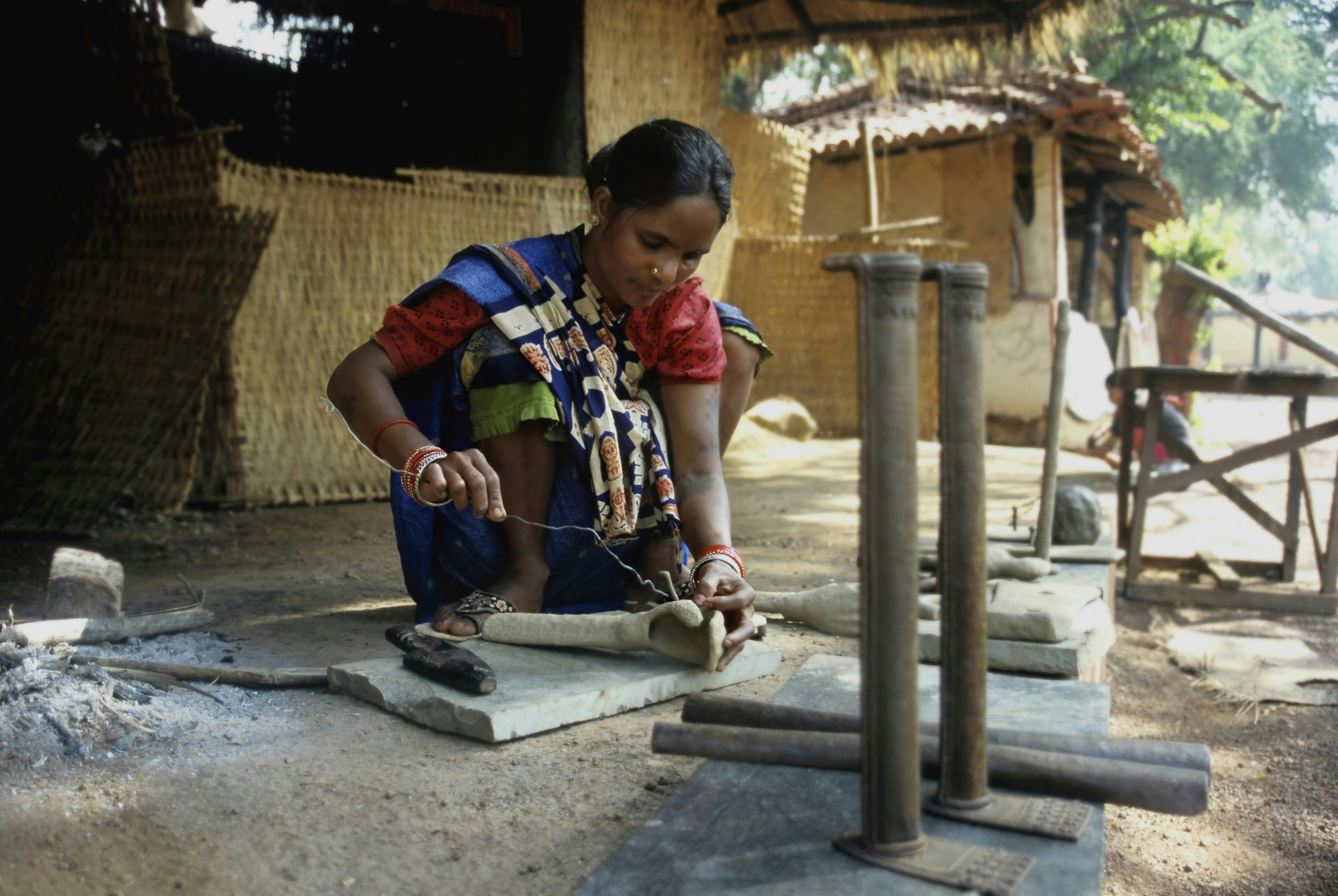 An Indian woman demonstrates the traditional art of dhokra casting, sculpting the arm of a statue using a thin piece of metal. The sculptor wears a brightly coloured sari while she works.