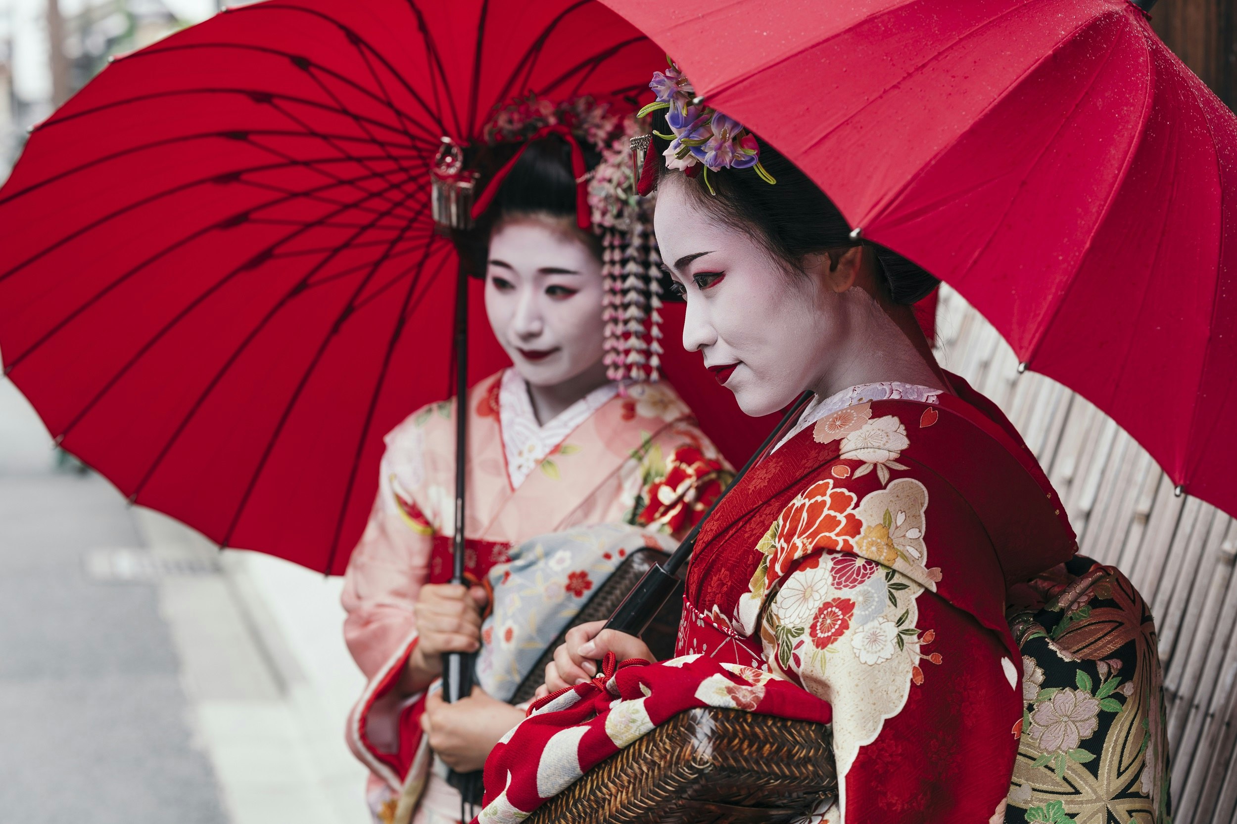 Two geisha in traditional attire stand beneath red umbrellas on the street in Gion.