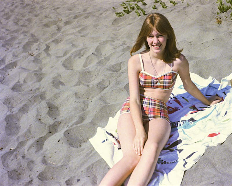 A girl sits on a sandy beach in a two-piece, checked bathing suit; she is looking up at the camera and smiling.
