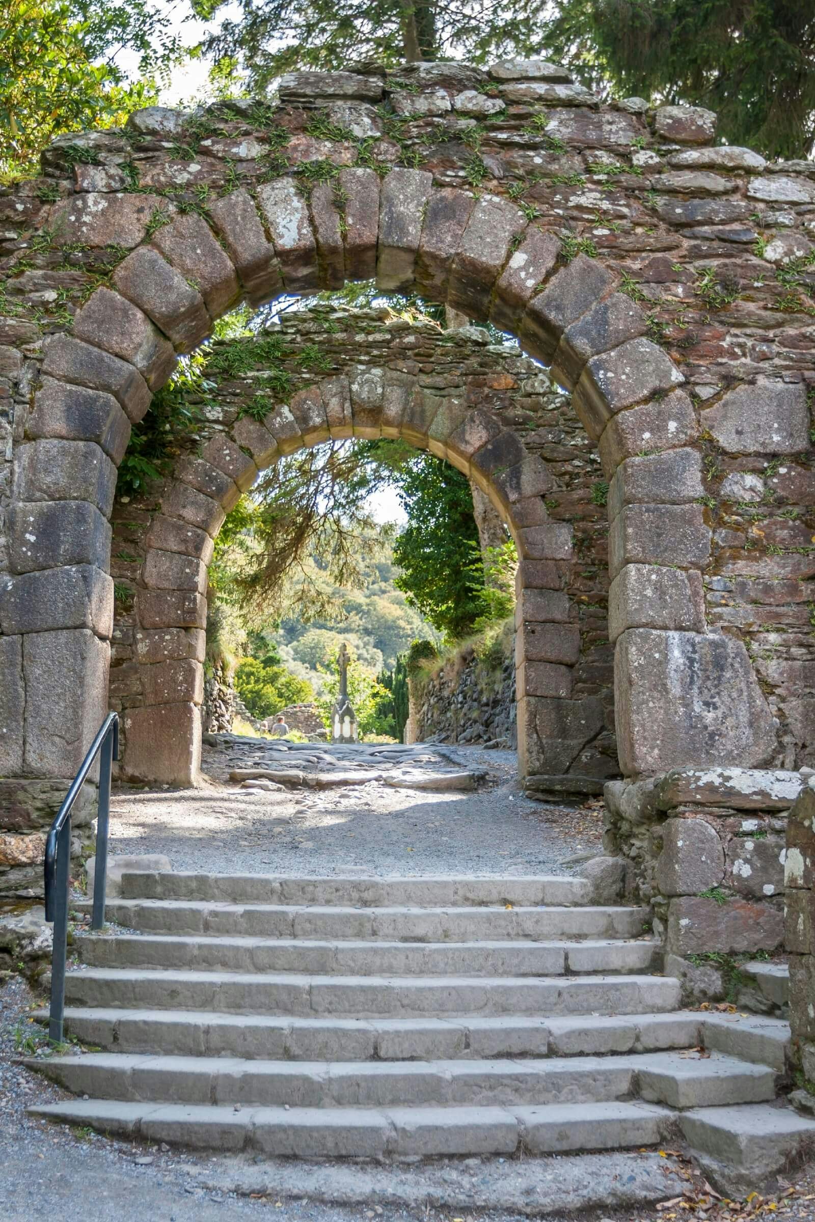 Steps going up to a stone arch with a forest and stone Celtic cross in the background