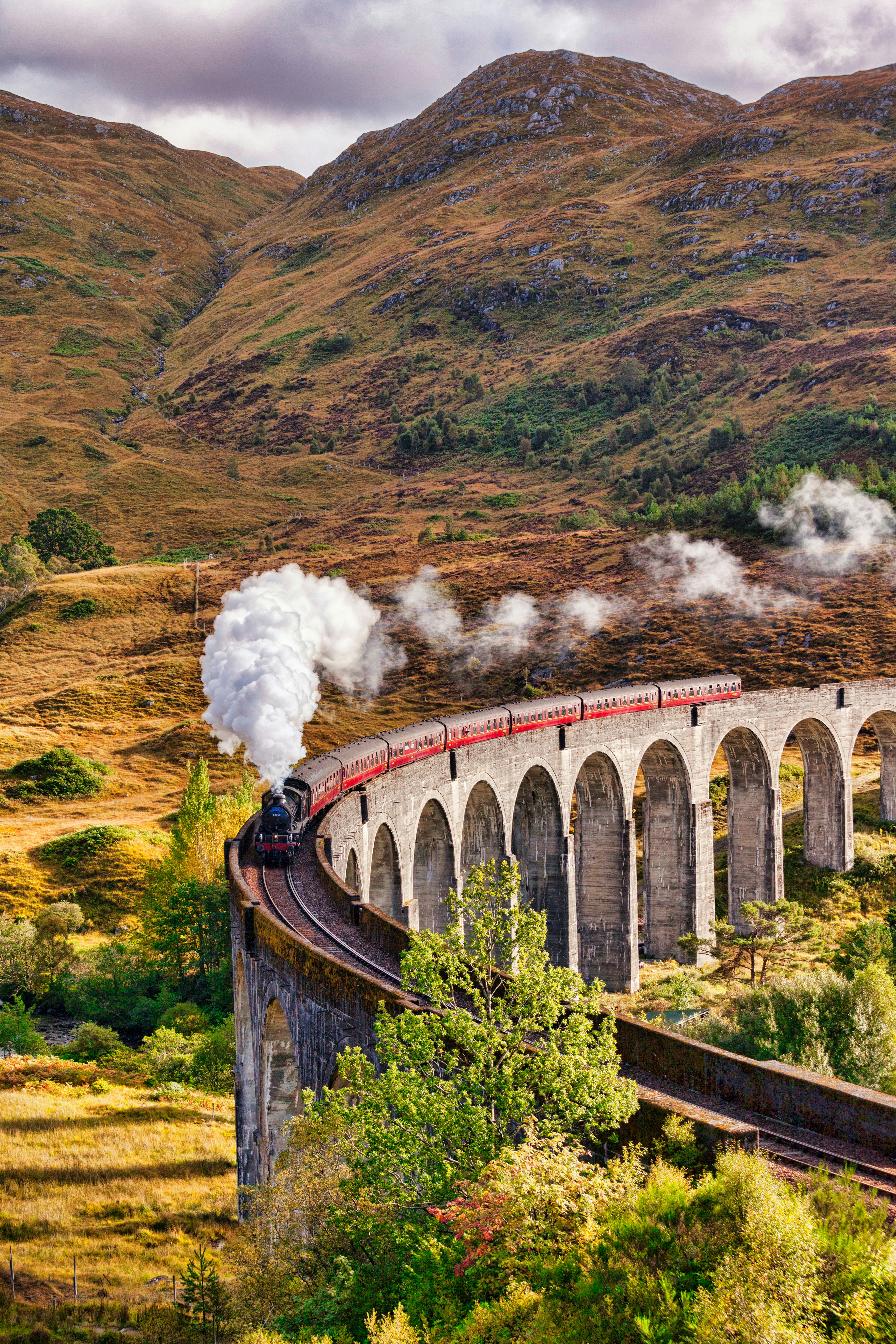 A steam train glides across Glenfinnan Viaduct; it's autumn and the landscape is painted with fall colours.