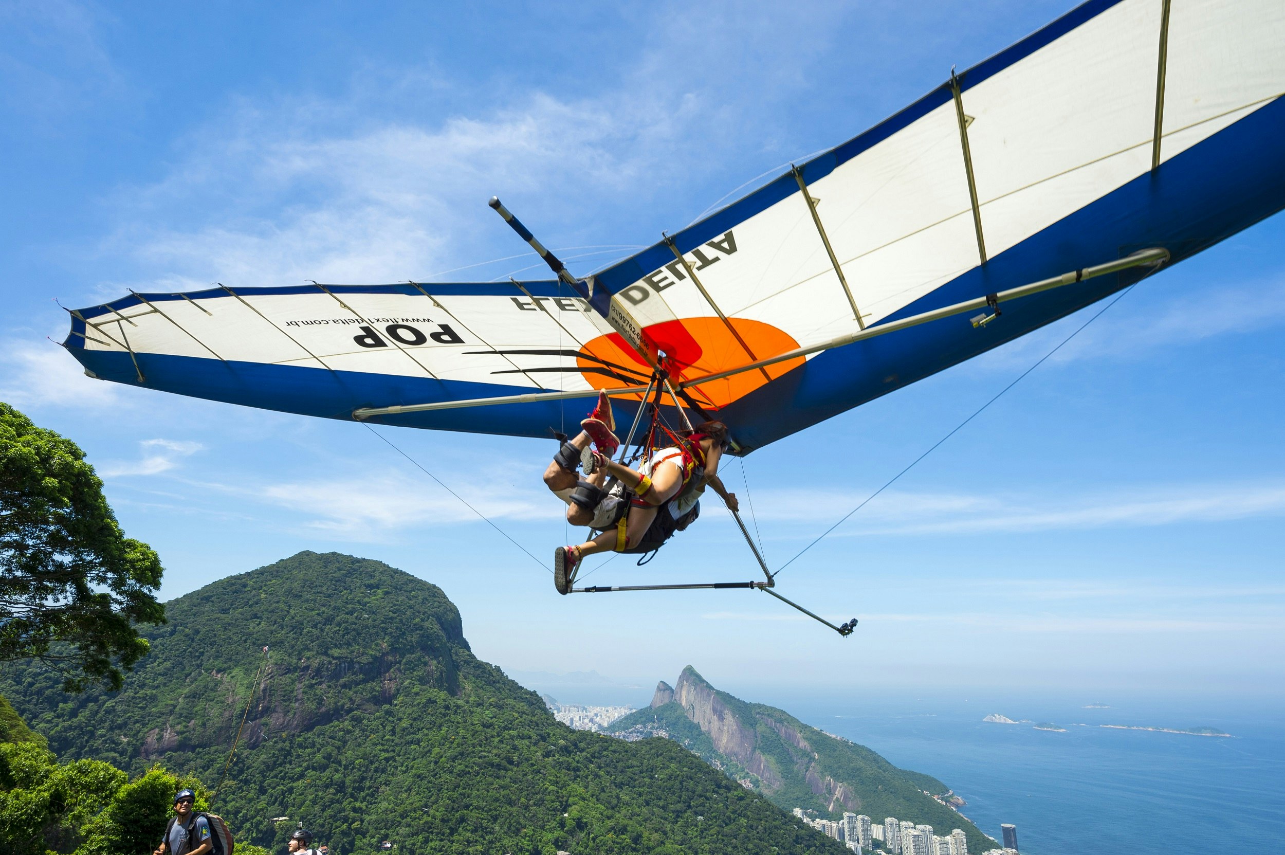 A hang gliding instructor takes off with a passenger from Pedra Bonita in the Tijuca National Forest; below them are forested hills and the distant buildings of Rio.