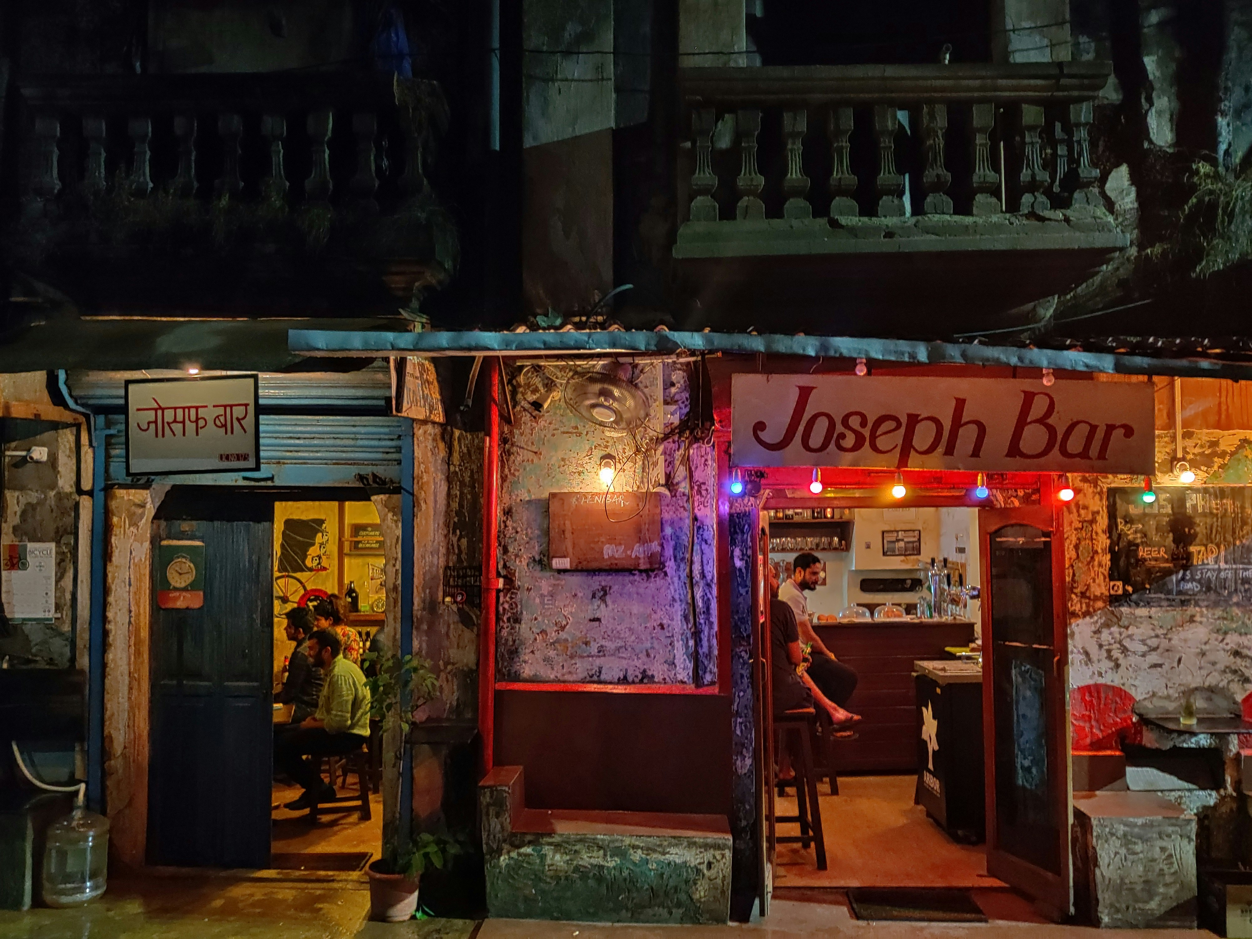 The dimly lit exterior of Joseph Bar in Goa is illuminated by red and purple string lights and the warm light pouring out of the bar itself. The entrance sits under a dark balcony, while inside a few patrons fill the doorways  