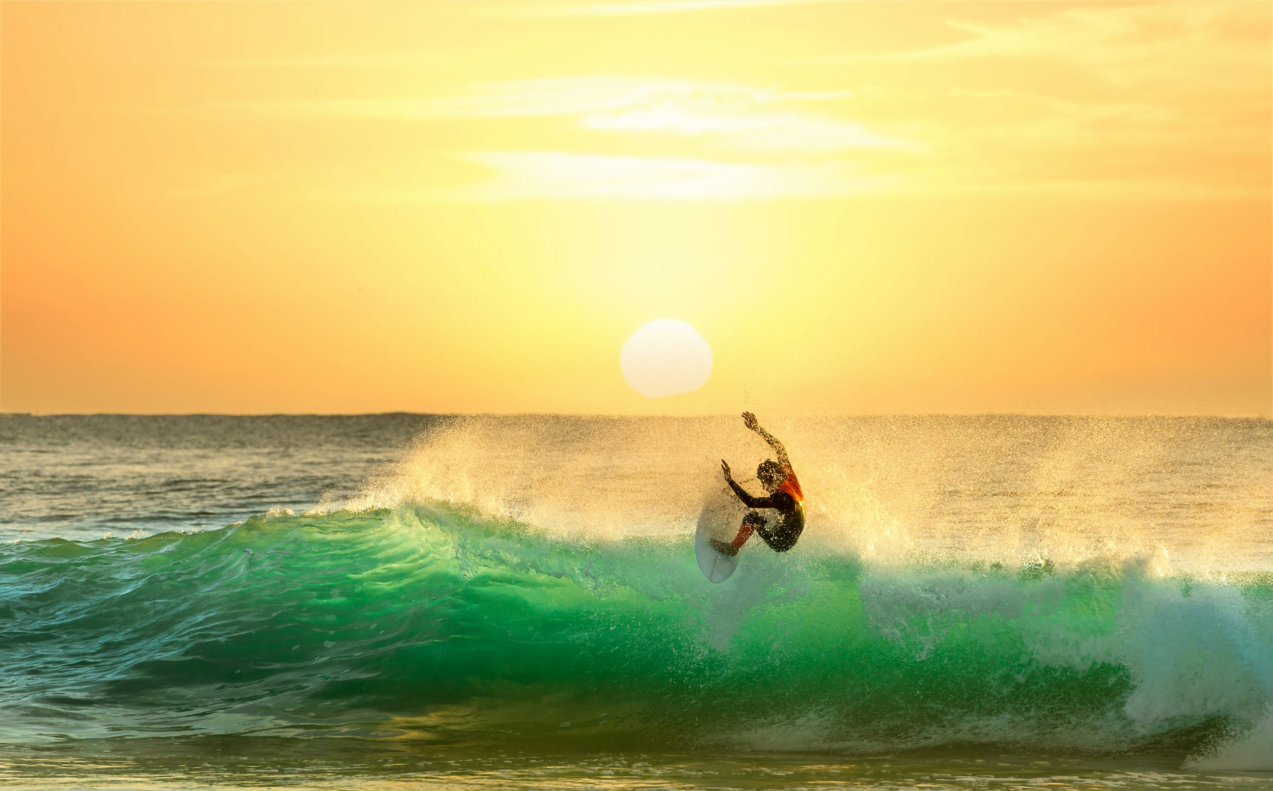 A surfer atop the crest of a wave almost floats in the air; the wave, lit by a sun right on the horizon, is glowing green. The sky is a brilliant gold colour.