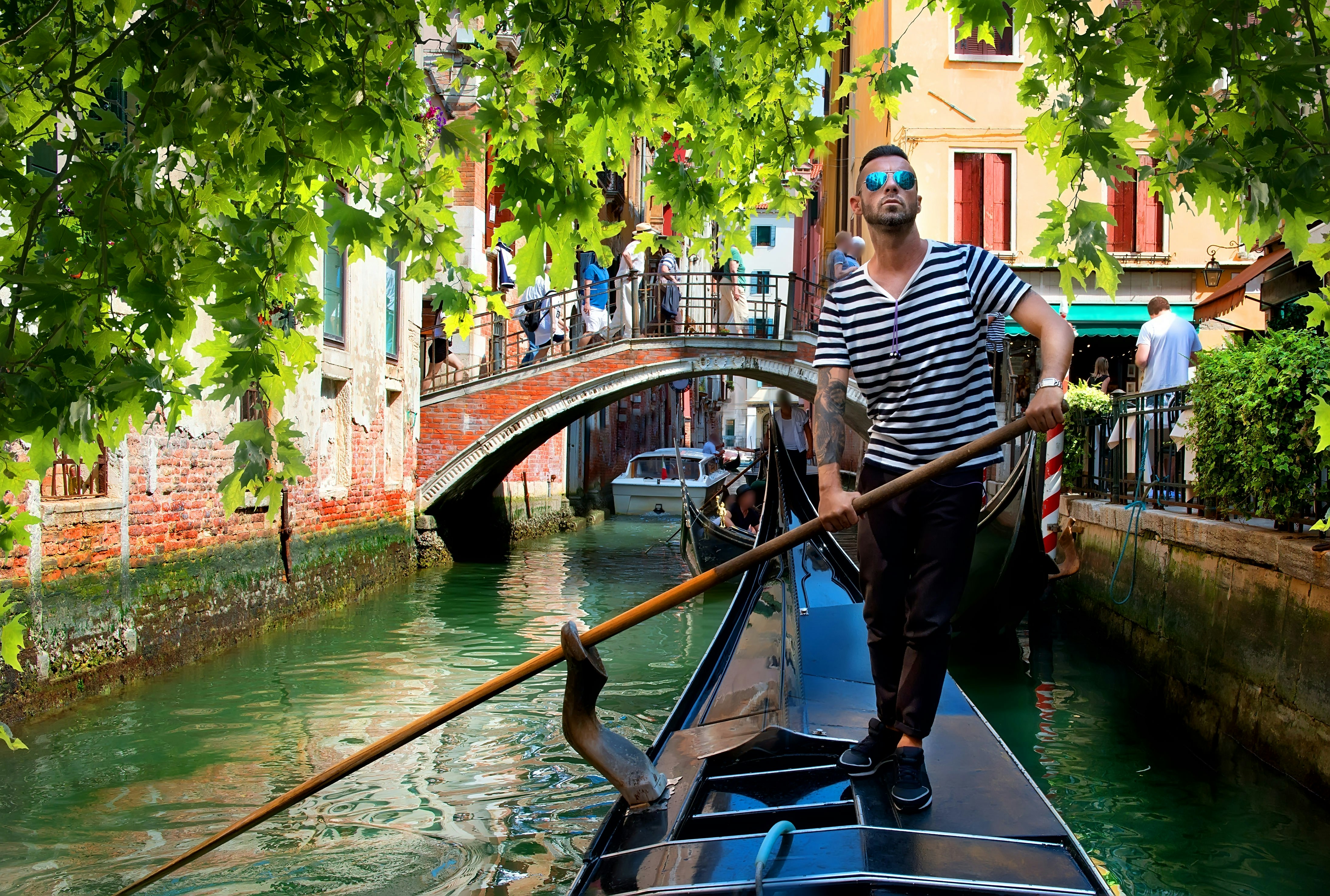 A male gondolier steers a boat through a narrow canal in Venice. The gondolier wears a black and white striped t-shirt, with black trousers. In the background, a number of people can be seen walking across a bridge that spans the canal.