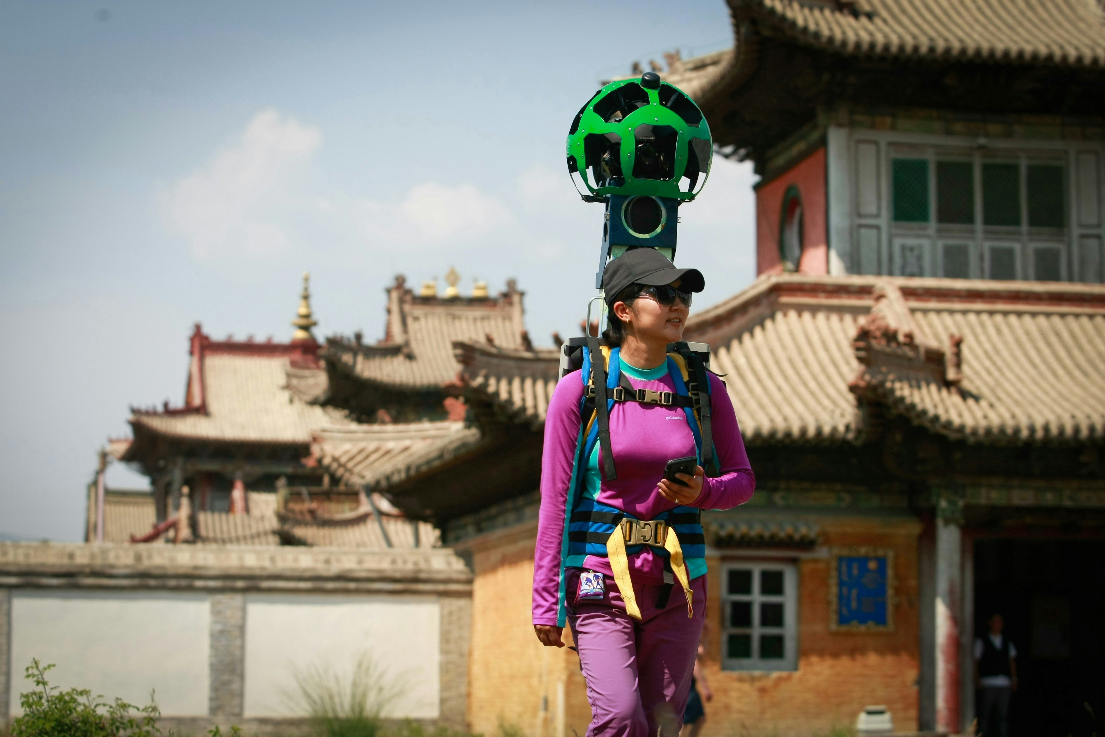 A woma nin a pink technical fabric top and matching pants and a black cap with a brim walks past the orange buildings and clay roofs of the Choijin Lama Museum in Ulan Bator with a green camera sphere from Google Street View strapped to her back