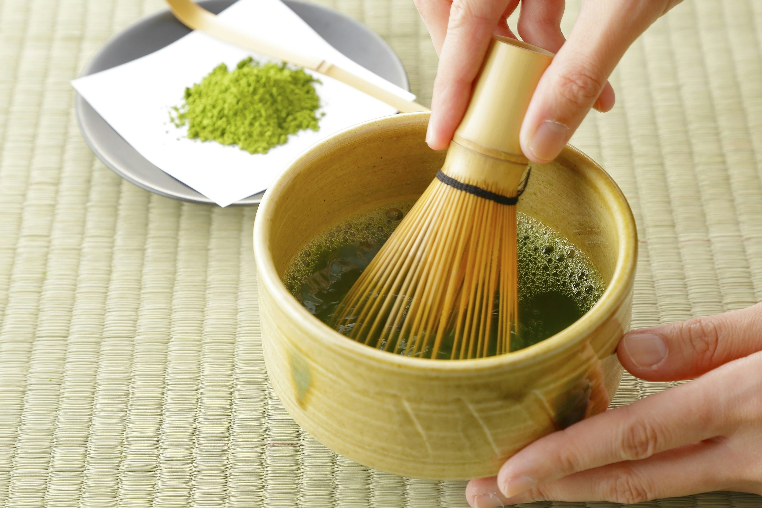A person whisks green tea in a small bowl with a bamboo utensil, with some powdered match sitting nearby on a small piece of paper on a plate.