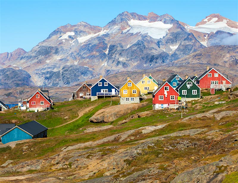 Greenland landscapes Colourful wooden houses dot a hillside. Snow-topped mountains rise up behind.