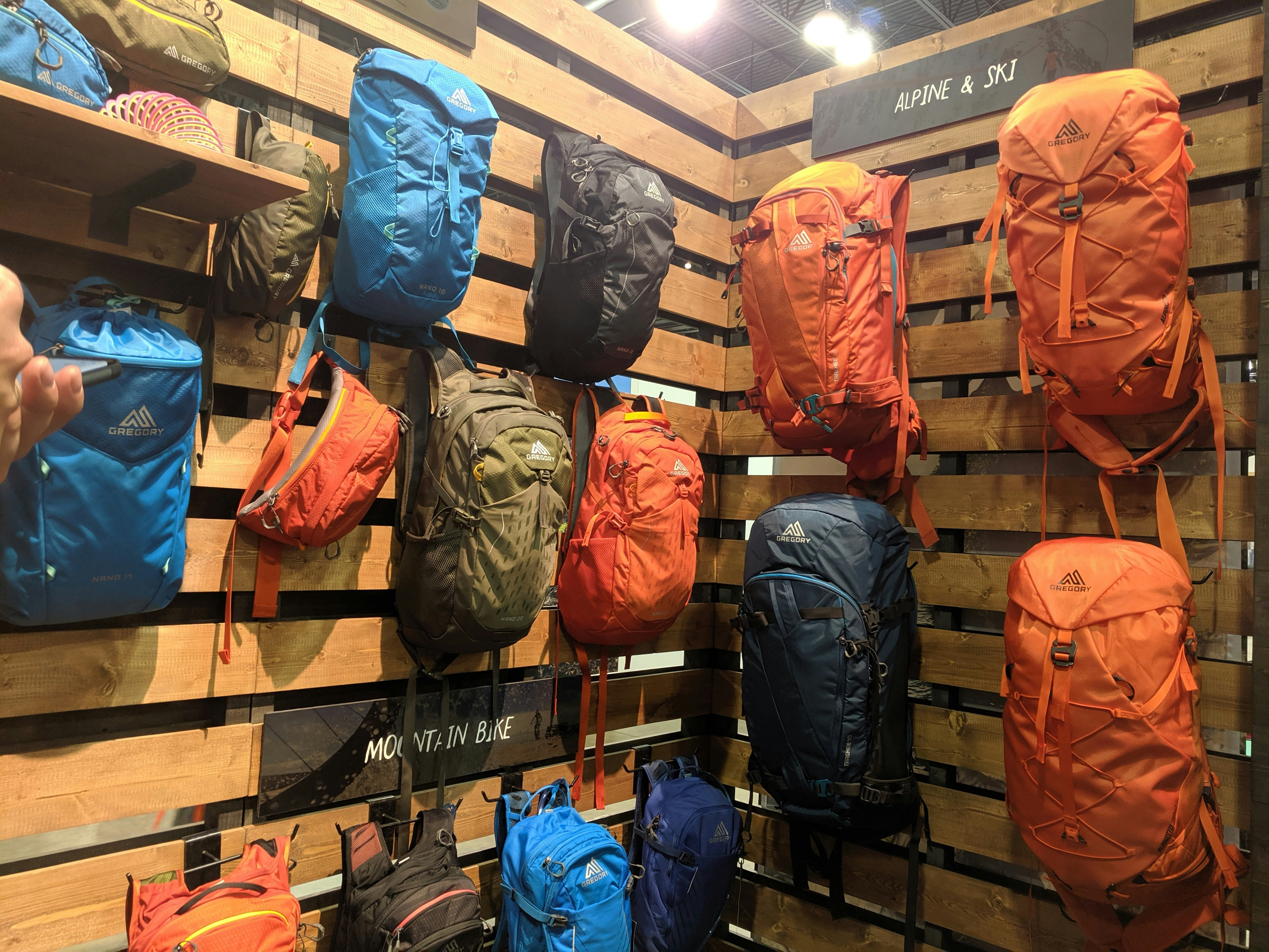 Brand new Gregory backpacks in orange, bright blue, olive, and black hang from a display wall made of warm wooden planks oriented horizontally at Outdoor Retailer Summer 2019
