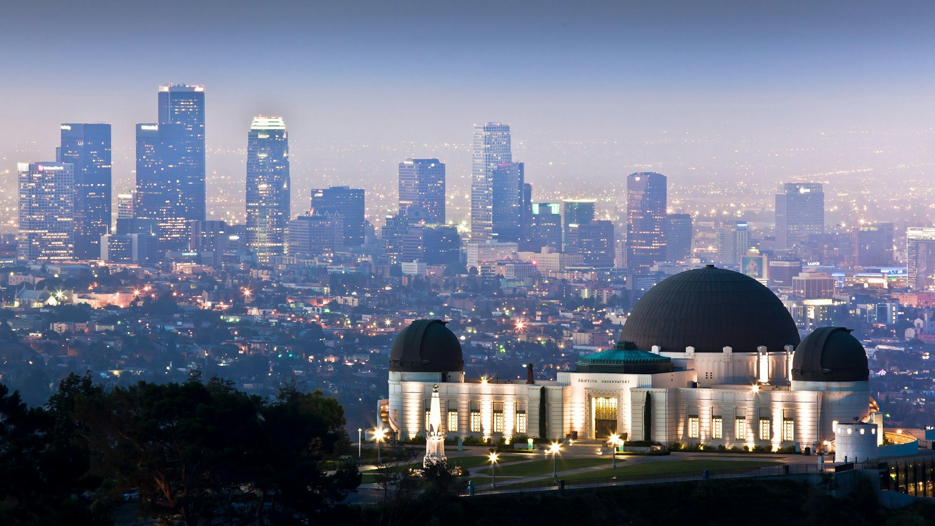 Shot from a distance, the art deco Griffith Observatory stands on the crest of a hill with the sparkling city skyline at dusk as its backdrop.