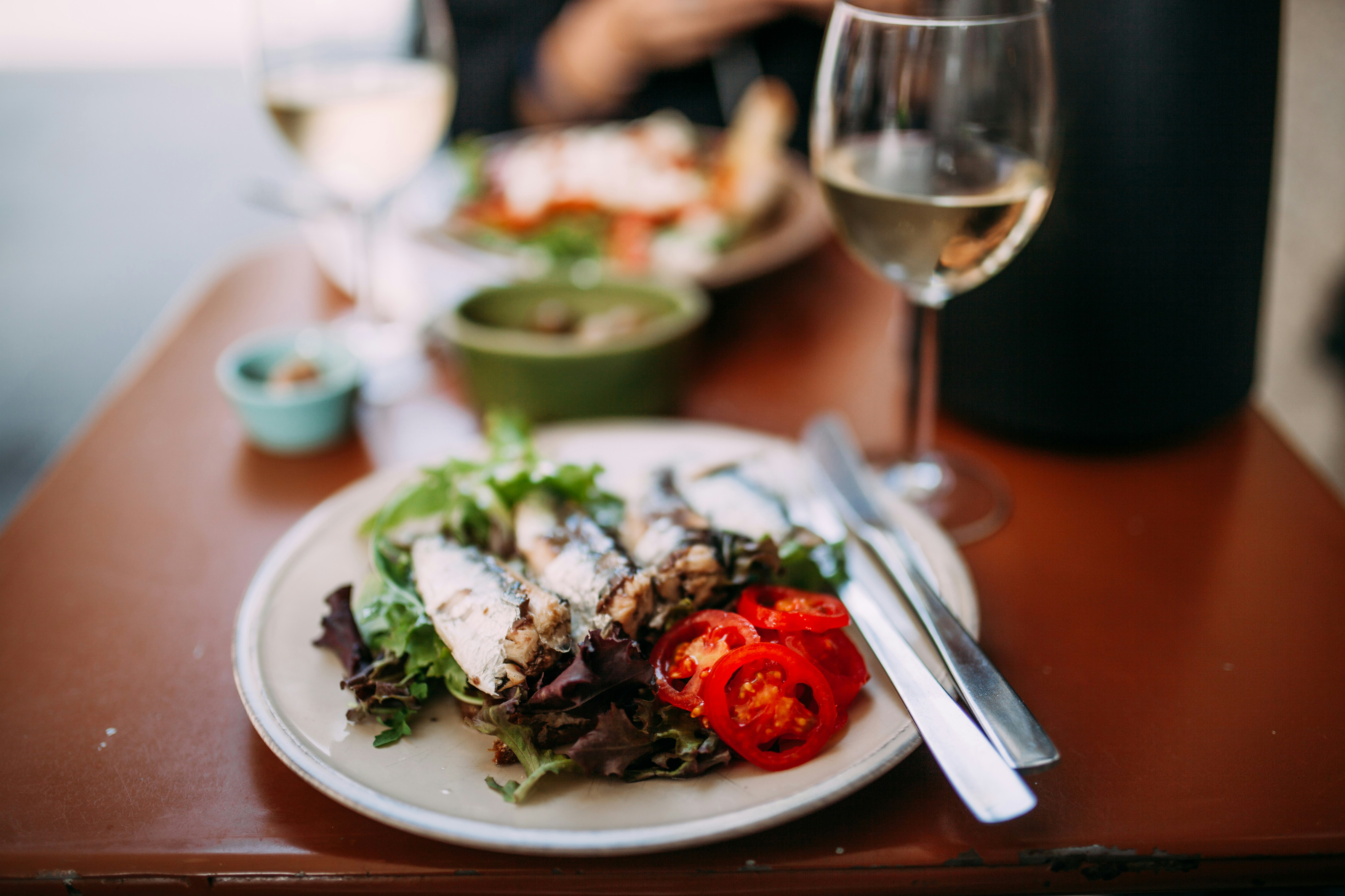 A plate sits in the foreground on a table, with three sardines sitting atop some mixed-leaf salad with sliced cherry tomatoes; in the background are blurred glasses of white wine.