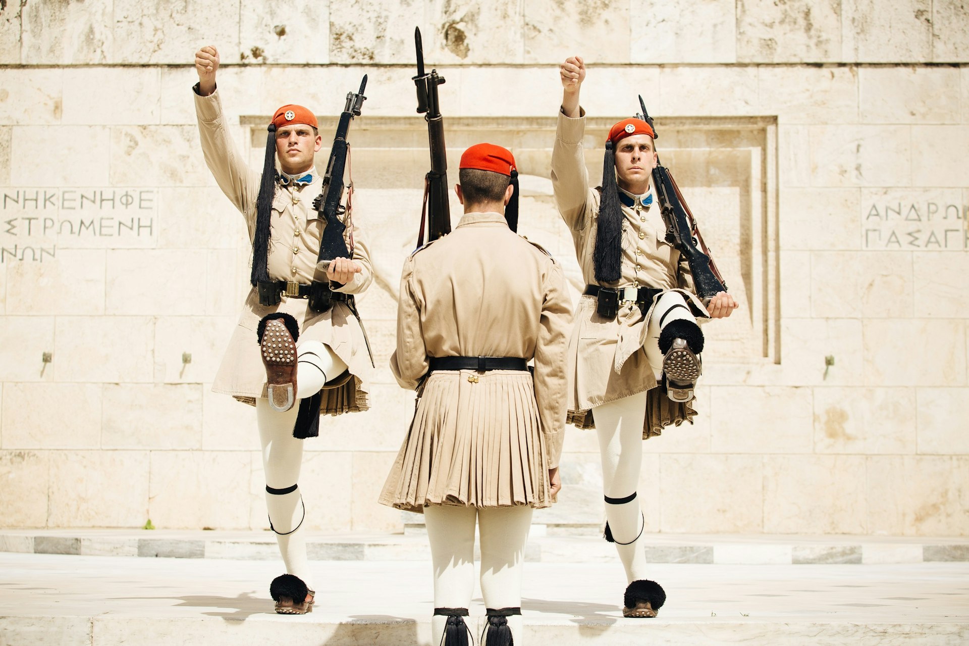 Two guards march towards the camera with their left legs kicked high; another guard has his back to the camera. All are wearing beige uniform with red caps and are armed with large rifles 