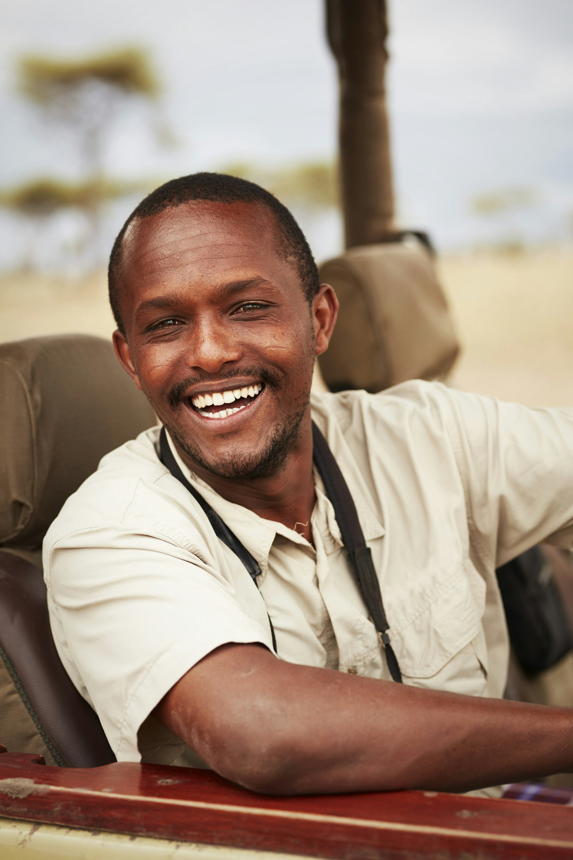 A smiling African safari guide sits in the driver's seat of an open-topped 4WD.