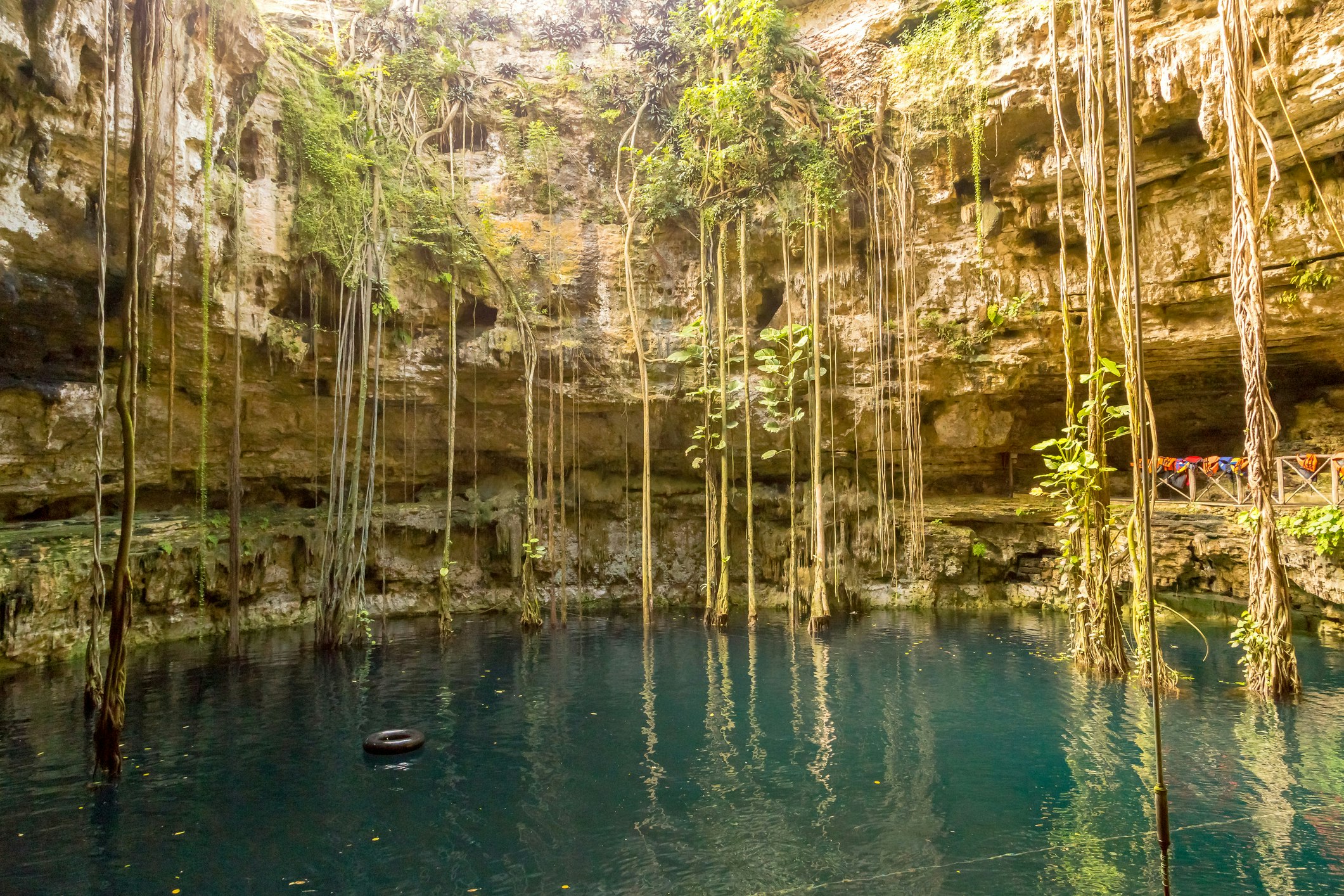 The deep blue waters of the cenote at Hacienda San Lorenzo are surrounded by buff and off-white stone walls strewn with long vines dangling from the top of the frame to the surface of the water. To the left, a black inner tube floats on the water.