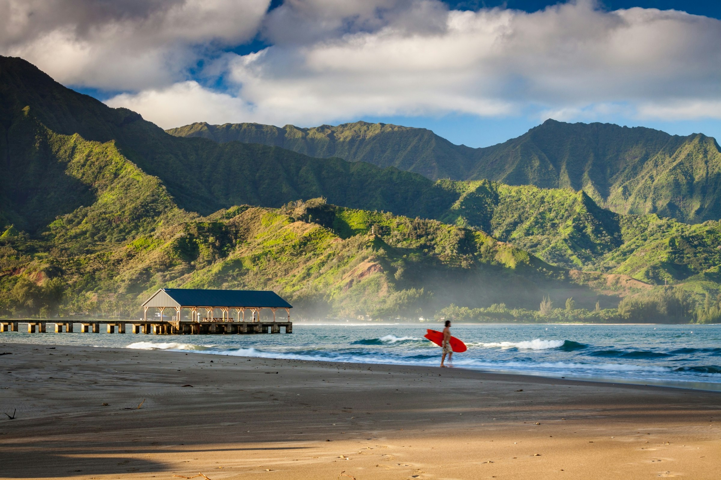 A lone surfer walks towards the water on the beach at Hanalei Bay, Hawaii.